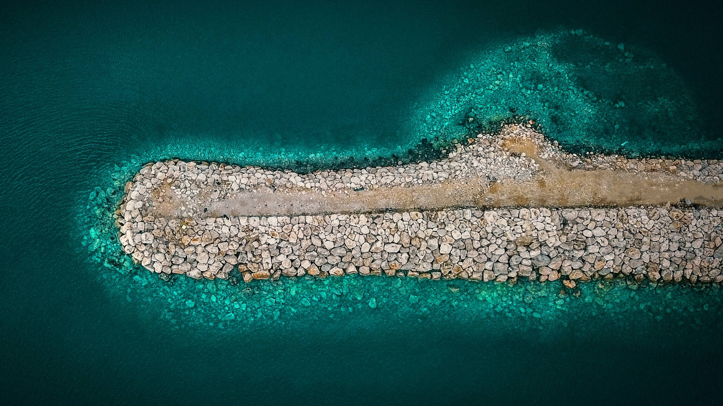 海洋藍色水域中的石板小徑 壁紙 — 石造防波堤延伸至充滿活力的綠松石色海水中的航拍視圖，邊緣清澈淺顯。