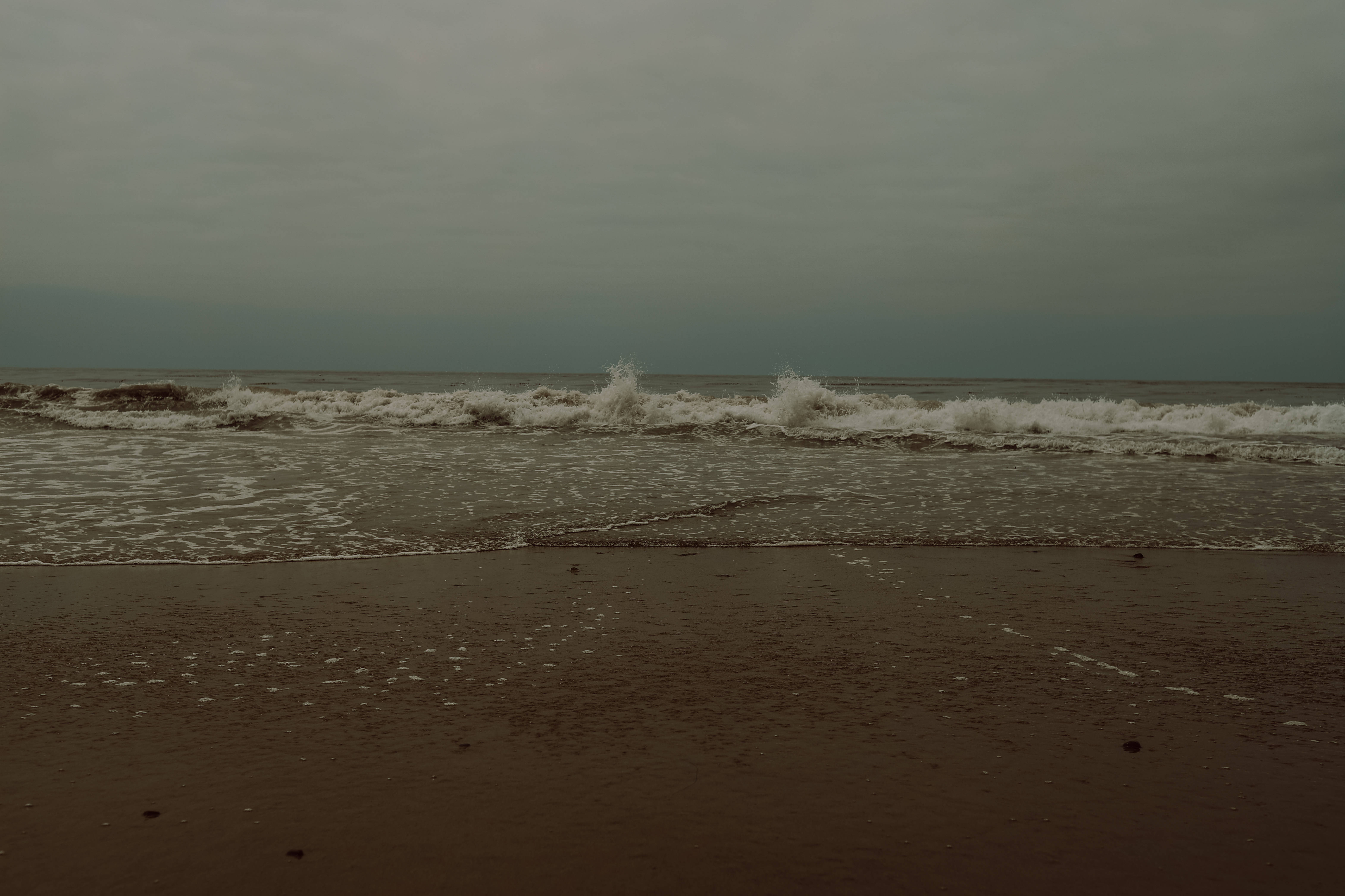 海浪意境筆電壁紙 壁紙 — 在厚重的陰天下，海浪拍打著潮濕的沙灘，呈現柔和的棕色和灰色色調。
