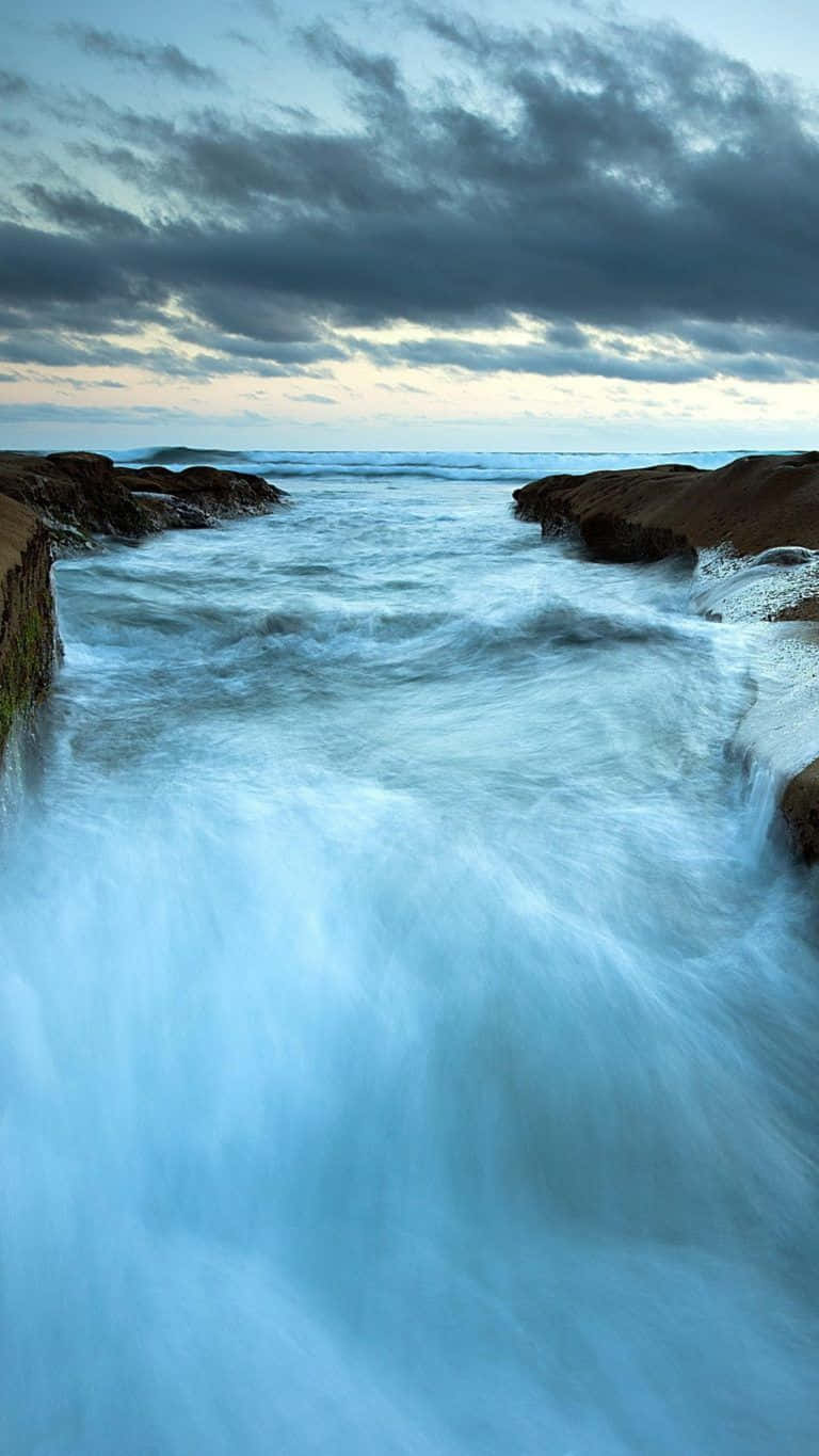 海浪拍打無聊的岩石 壁紙 — 強勁的海浪衝擊崎嶇的懸崖，天空陰沉，暮色中雲層條紋分明。