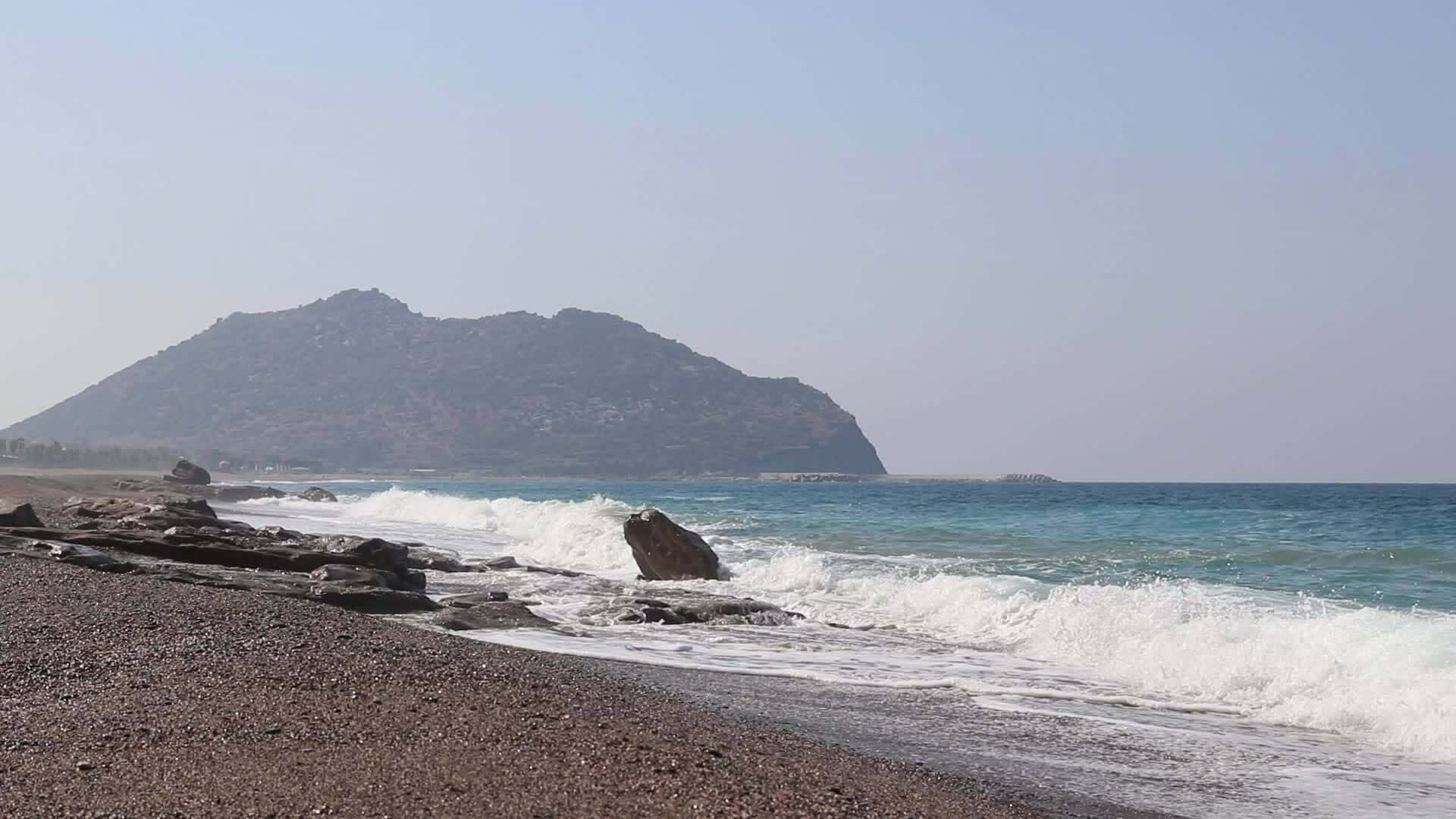 Mediterranean Beach 桌布 壁紙 — 海浪拍打著陽光普照的海岸上的深色鵝卵石，崎嶇的山巒在朦朧的遠方升起。