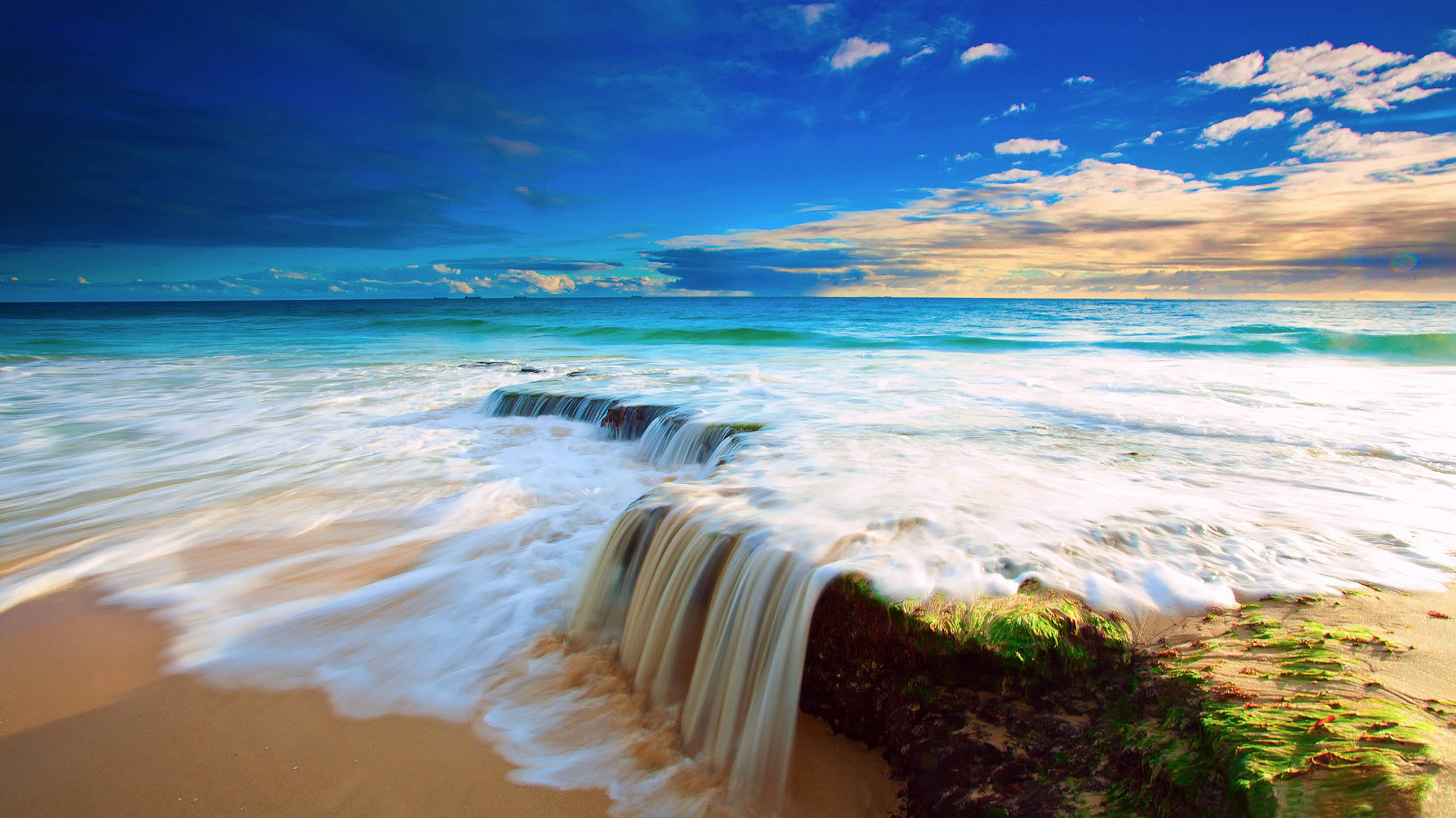 海浪瀑布 壁紙 — 海浪越過岩石防波堤，傾瀉在金色沙灘上，天空湛藍，白雲點綴。