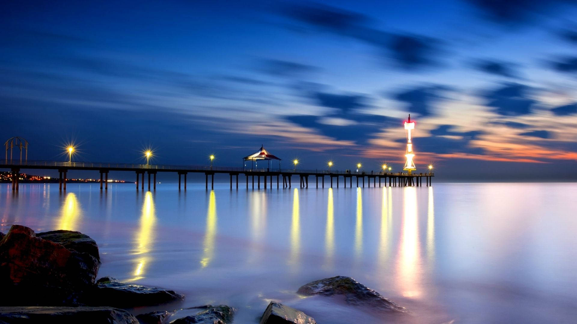 海濱夜景天際 壁紙 — 一條長長的碼頭延伸至平靜的水面，在暮色中，發光的燈光映照在水面上，天空呈現條紋狀的藍色和橙色。