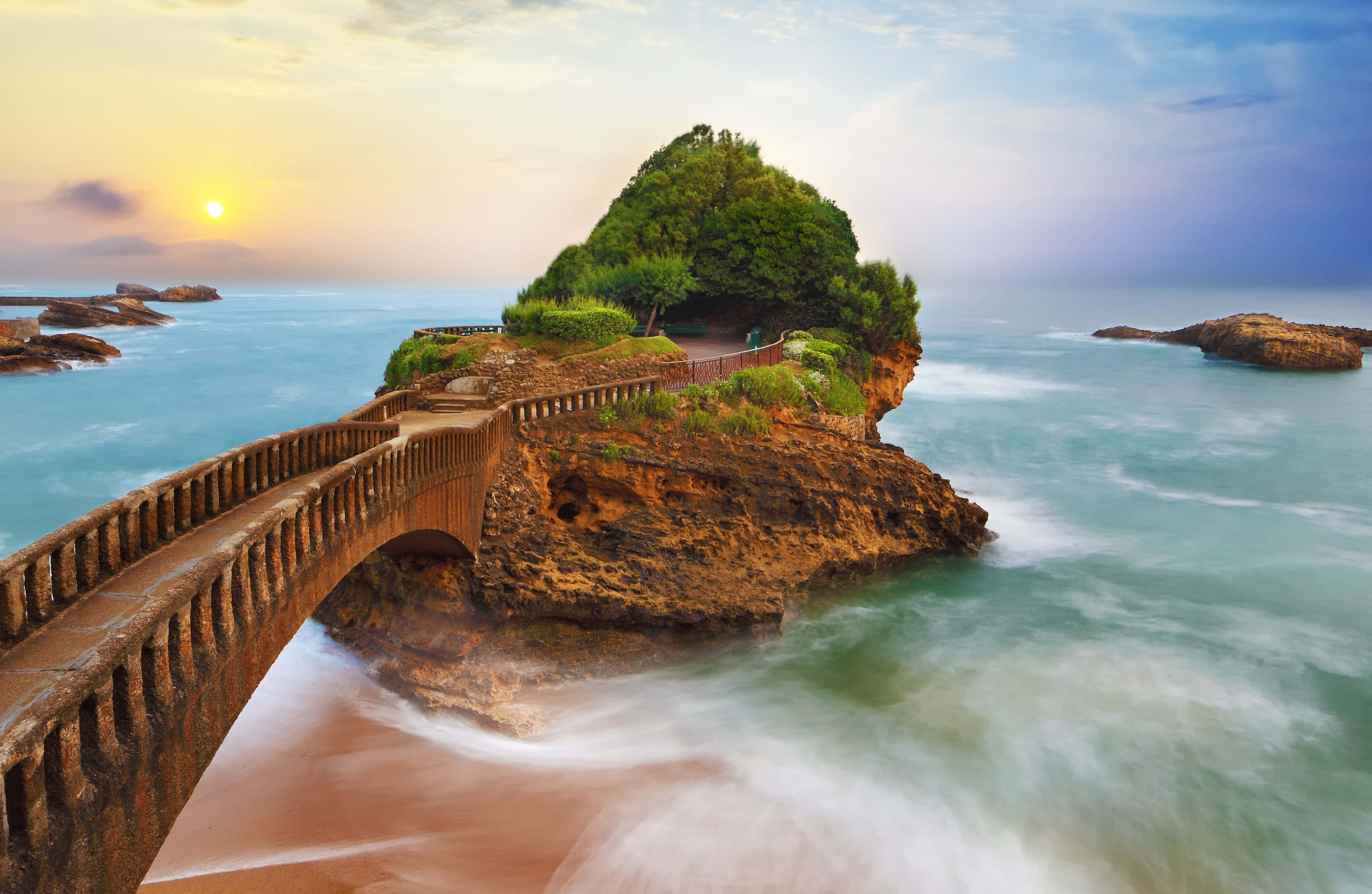 海濱風景 壁紙 — 一座石橋拱起在碧綠色的水上，通往一個在日落時分被柔和金色光芒籠罩的鬱鬱蔥蔥的綠色島嶼。