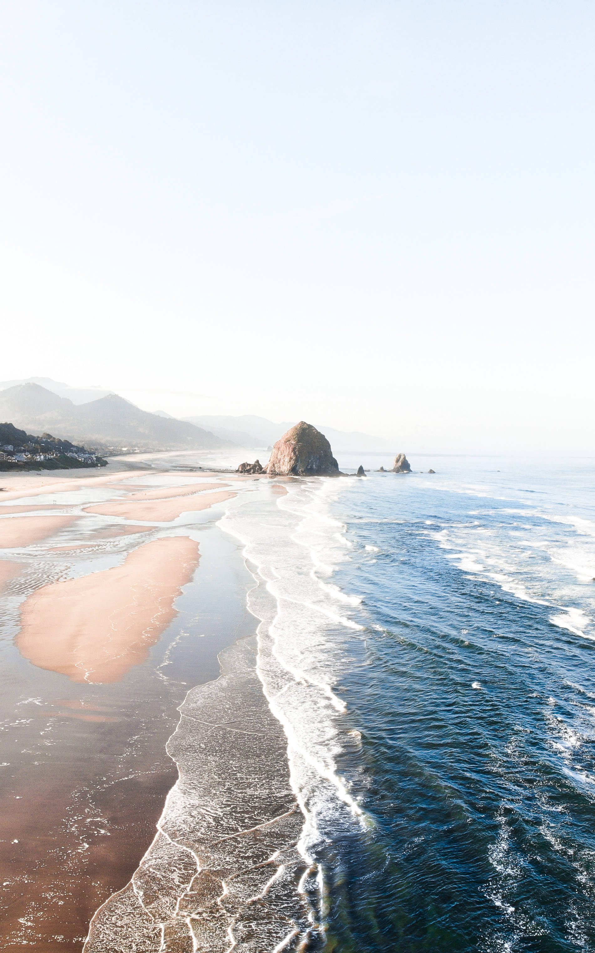 海灘 iPhone 壁紙：海灘岩 壁紙 — 在朦朧的天空下，一個寧靜的海岸場景，溫柔的海浪拍打著粉紅色的沙灘，旁邊有一個巨大的海蝕柱。