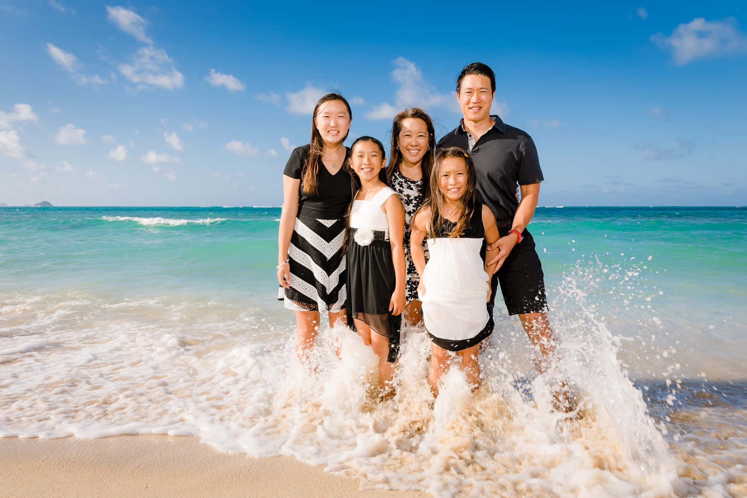 海灘上完美的一天，全家共享幸福時光！ 壁紙 — 一個快樂的五口之家站在淺海水中，海浪在他們腳邊飛濺，頭頂是明亮的藍天。