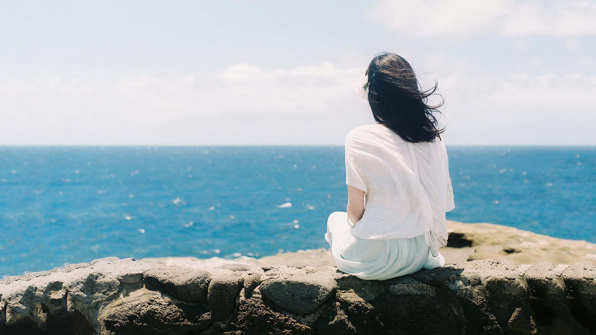 身穿白衣的女子坐在石牆上，面向日落時分的海洋