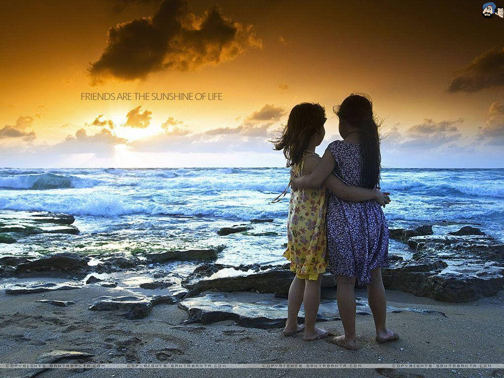 海灘上的女孩友誼 壁紙 — 兩個女孩在岩石海灘上擁抱，金色夕陽的光芒沐浴著她們身後的海洋波浪。