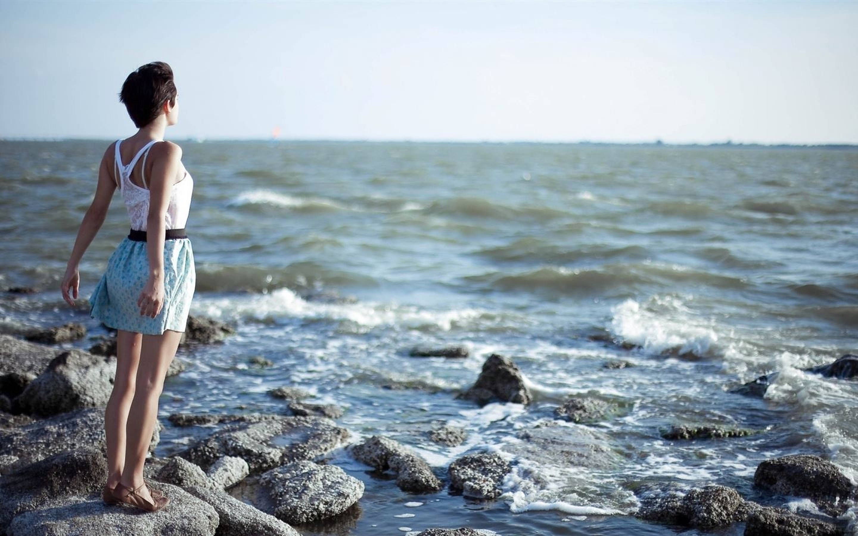 海灘上的女性呼吸 壁紙 — 一名女子赤腳站在岩石海岸上，在明亮的日光下凝視著廣闊的海洋。