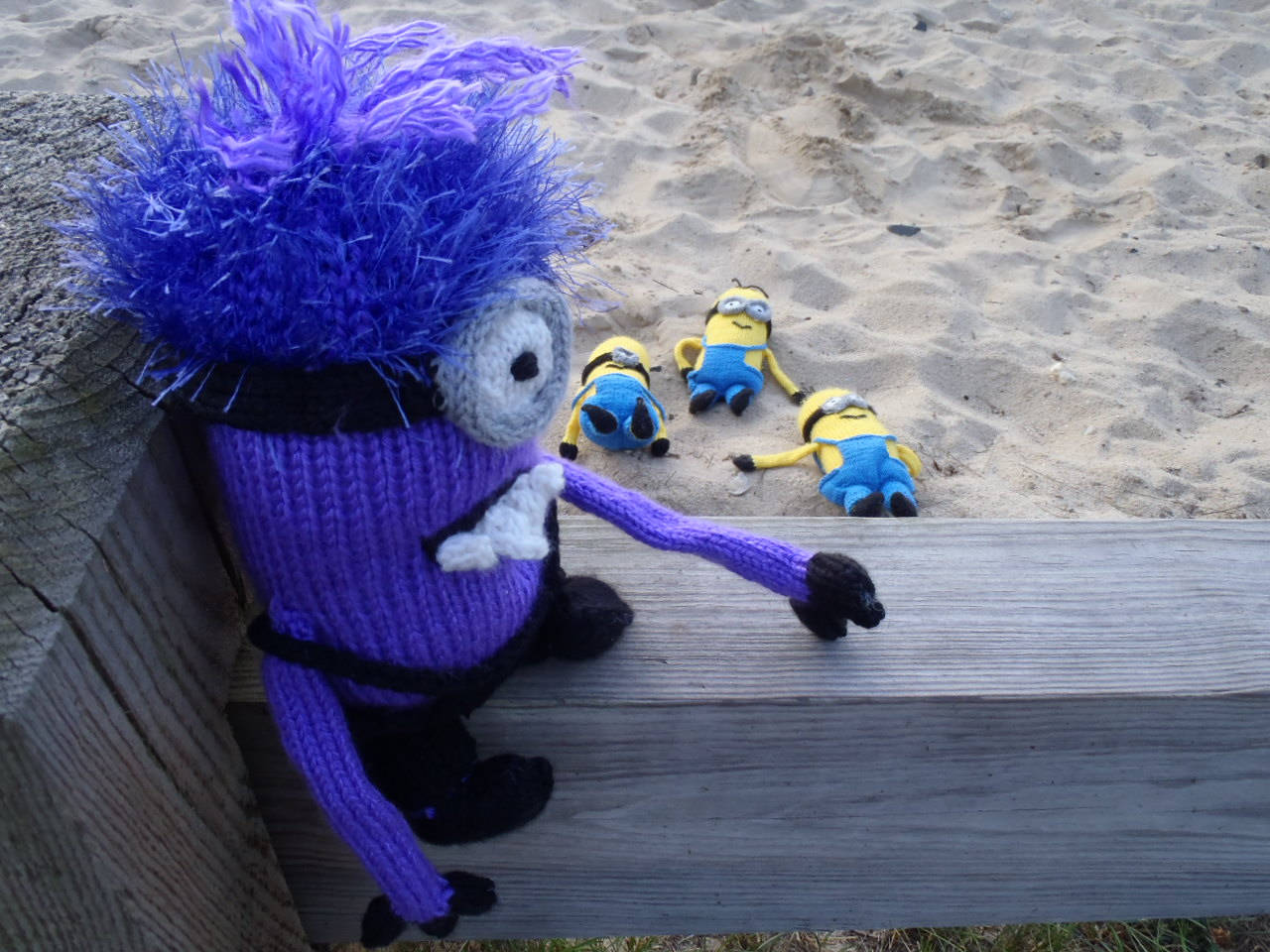 海灘上的惡魔小兵 壁紙 — 一個編織的紫色Minion，頭髮凌亂，靠在木欄杆上，而三個黃色Minion在後面的沙灘上睡覺。