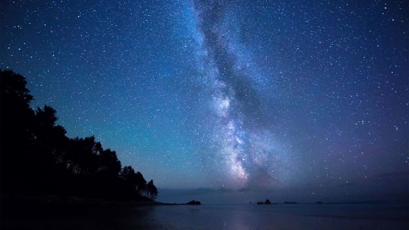 海灘上的星空筆記本電腦壁紙 壁紙 — 一個令人屏息的夜空，充滿無數星星和發光的銀河，在平靜的海洋上空拱起，岸邊有黑暗的樹木剪影。