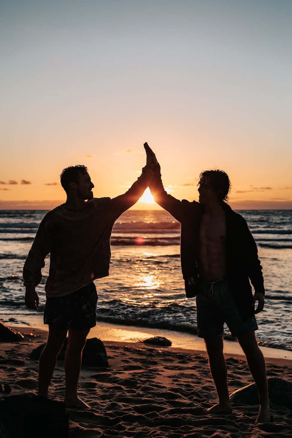 海灘上的美好友誼 壁紙 — 兩個剪影男人在沙灘上擊掌，夕陽在他們身後落下，在海面上投射出金色的光芒。