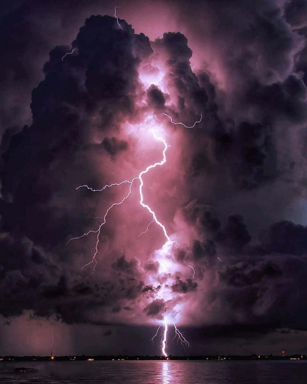 海灘上的美麗暴風雨天氣 壁紙 — 一道強勁的紫色閃電劃破暴風雨的天空，映照在黑暗的水面上，反射出遠處的城市燈光。