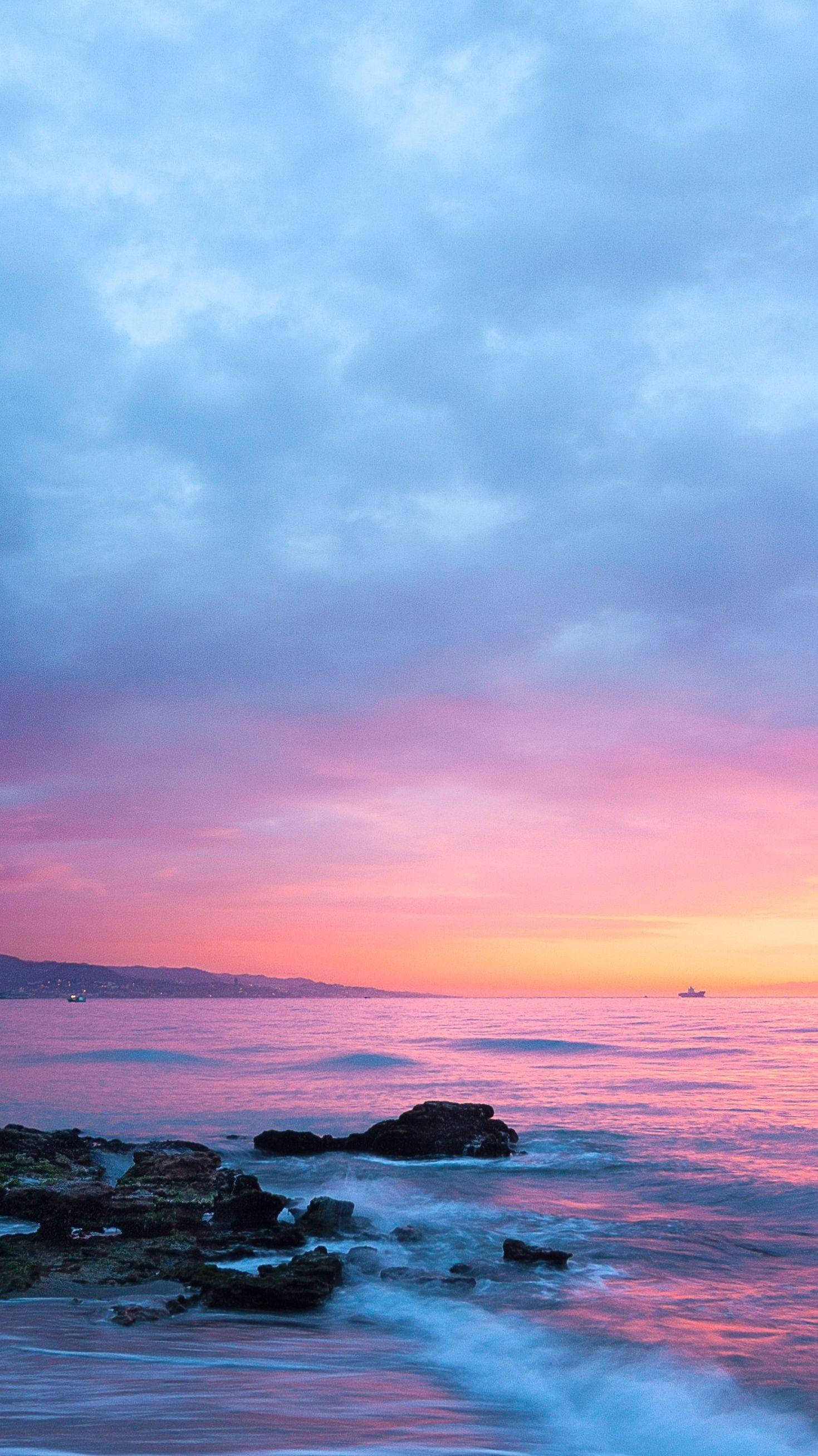 海灘夕照氛圍 壁紙 — 日落時分寧靜的海洋，粉紅、橙和藍色調映照在平靜的水面和前景的深色岩石上。