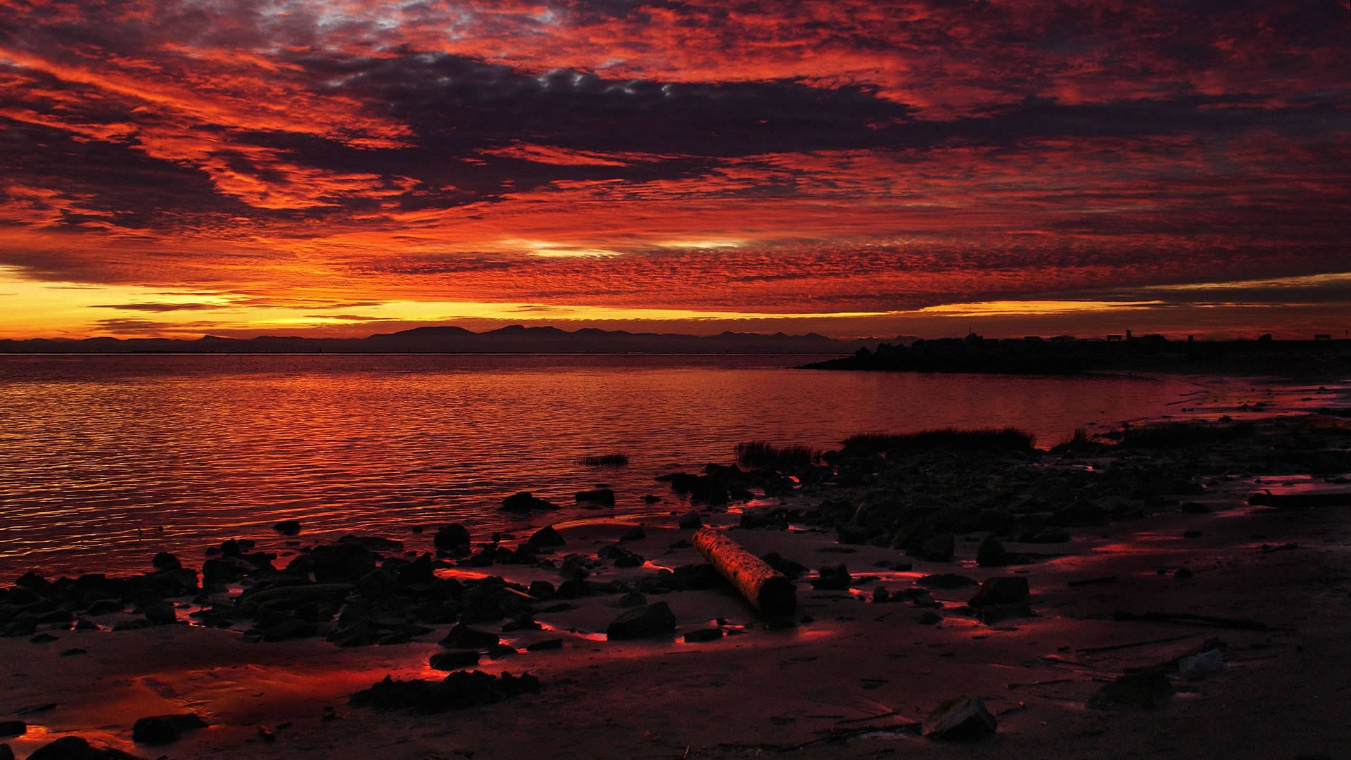 海灘夕陽風景 壁紙 — 熾熱的夕陽將天空和水面染成深紅和橙色，雲朵在岩石海岸線上發光。