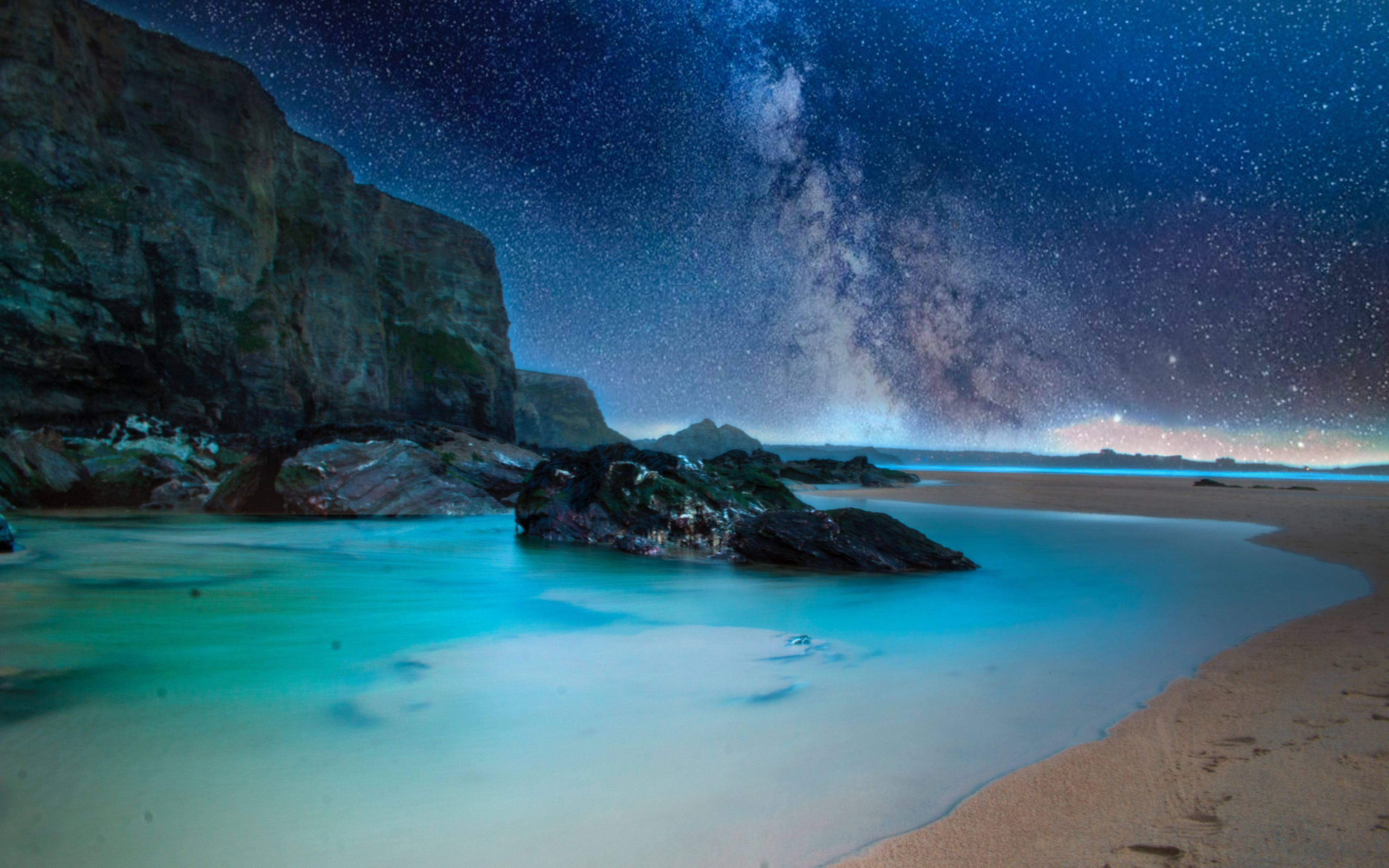 海灘夜景中的冷峻岩層 壁紙 — 銀河在寧靜的夜晚海灘上空拱起，藍綠色的水流過黑暗的岩石和懸崖。