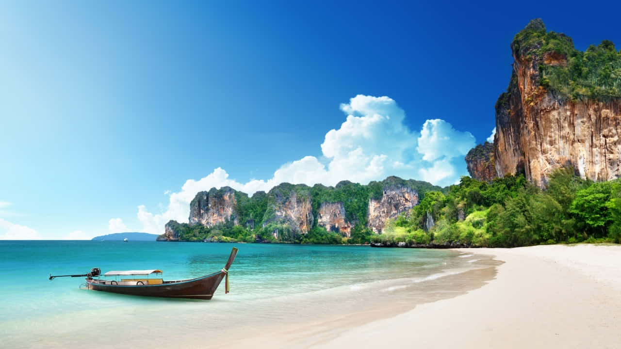 海灘寧靜日景 壁紙 — 一艘長尾船停泊在 pristine 白色沙灘上，旁邊是碧綠色的海水，背後是戲劇性的石灰岩懸崖，在茂密的綠植和明亮的藍天下聳立。