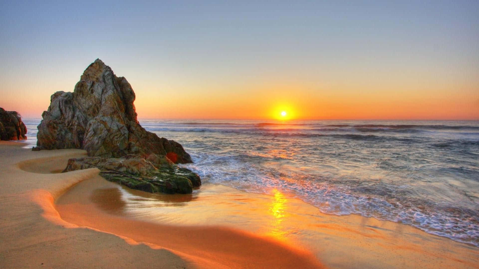 海灘岩石日落桌面 壁紙 — 夕陽將海洋和沙灘染成溫暖的橙色色調，海浪輕輕拍打著一個巨大的岩石突出物。