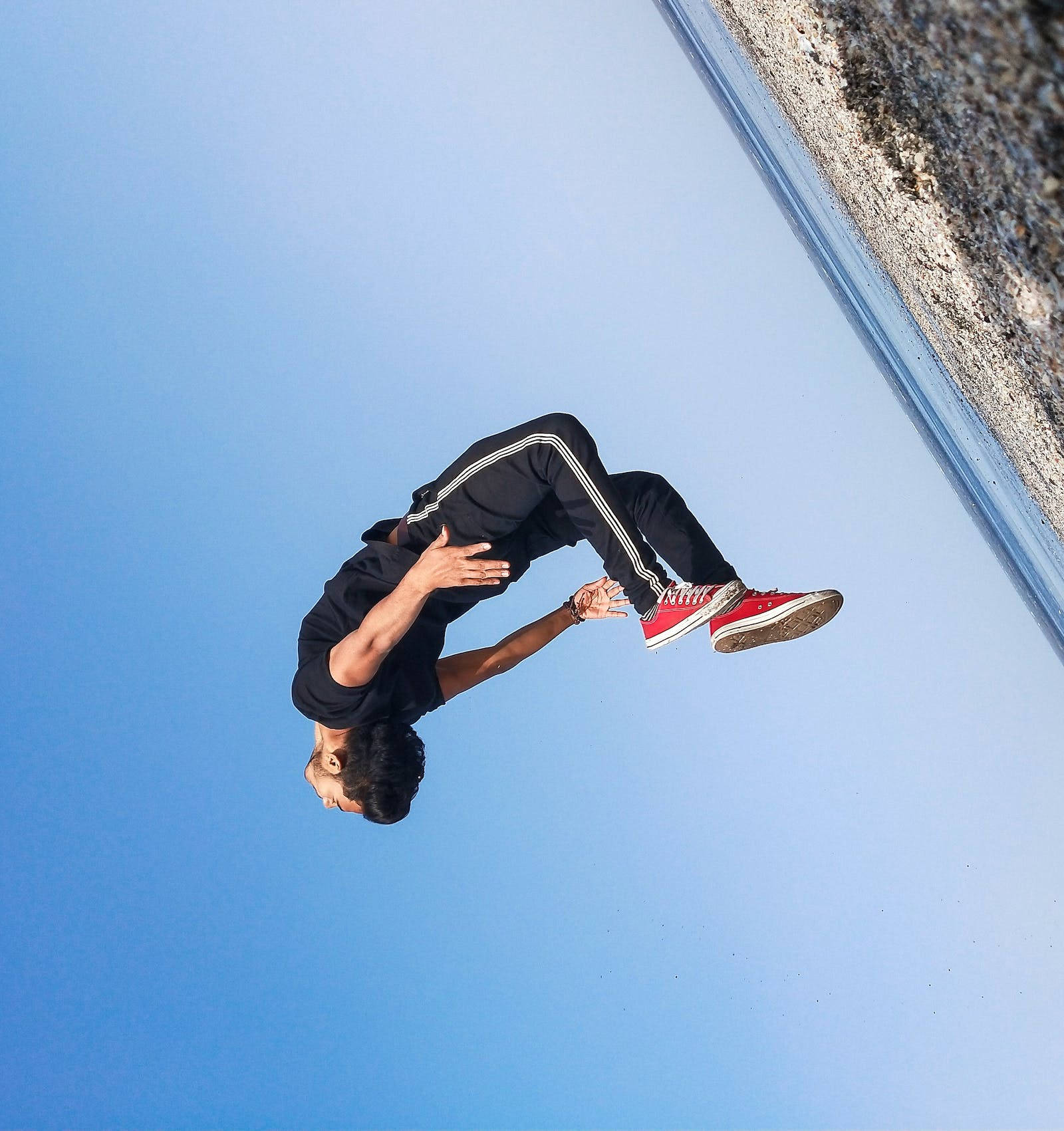 海灘後空翻跳躍 壁紙 — 一名身穿黑色運動服和紅色運動鞋的年輕男子，在岩石海岸線附近清澈的藍天下做出動態的空中翻騰動作。