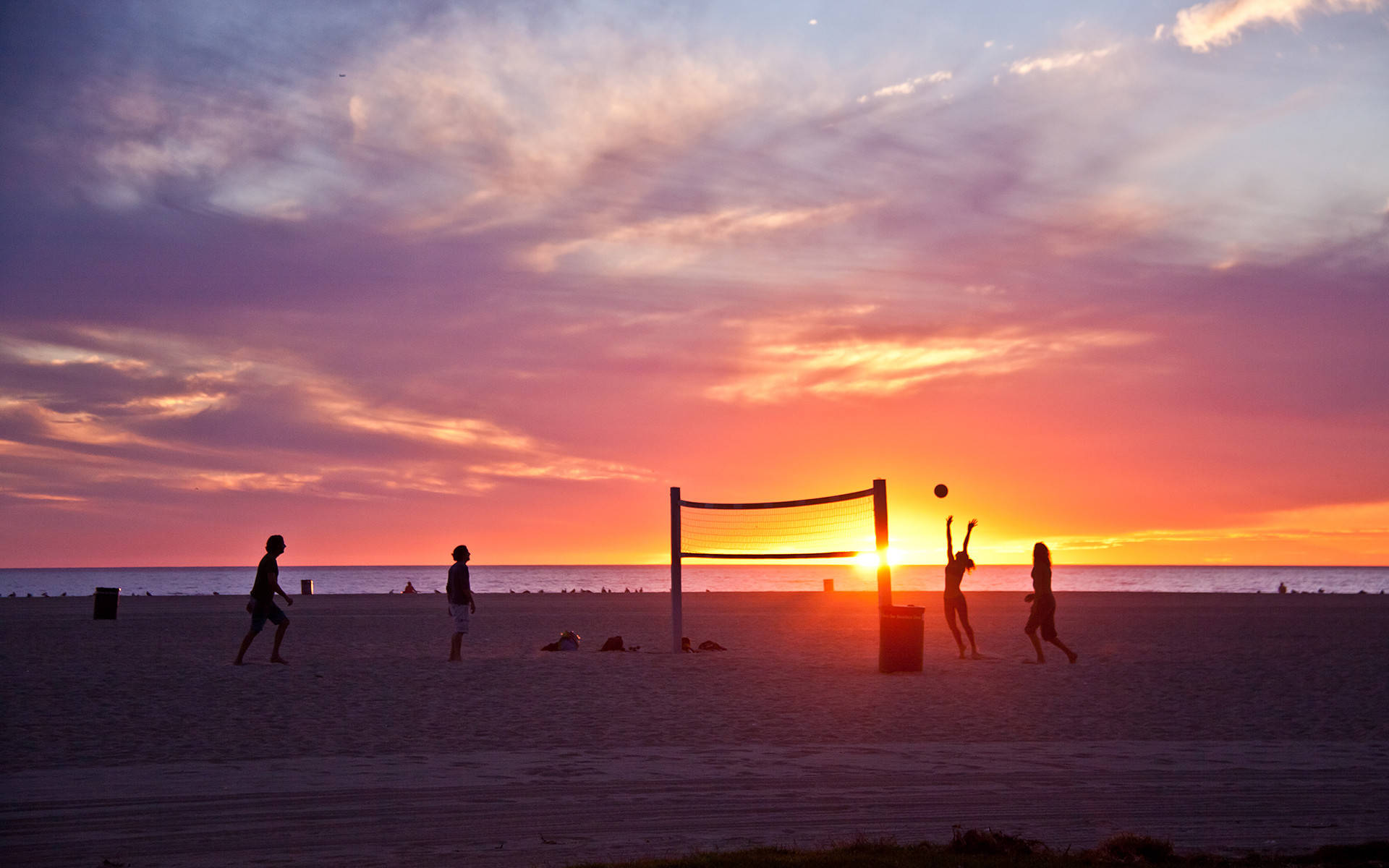 海灘排球與夕陽剪影 壁紙 — 剪影的沙灘排球運動員在海洋上空火熱的日落天空下跳躍和閃避。