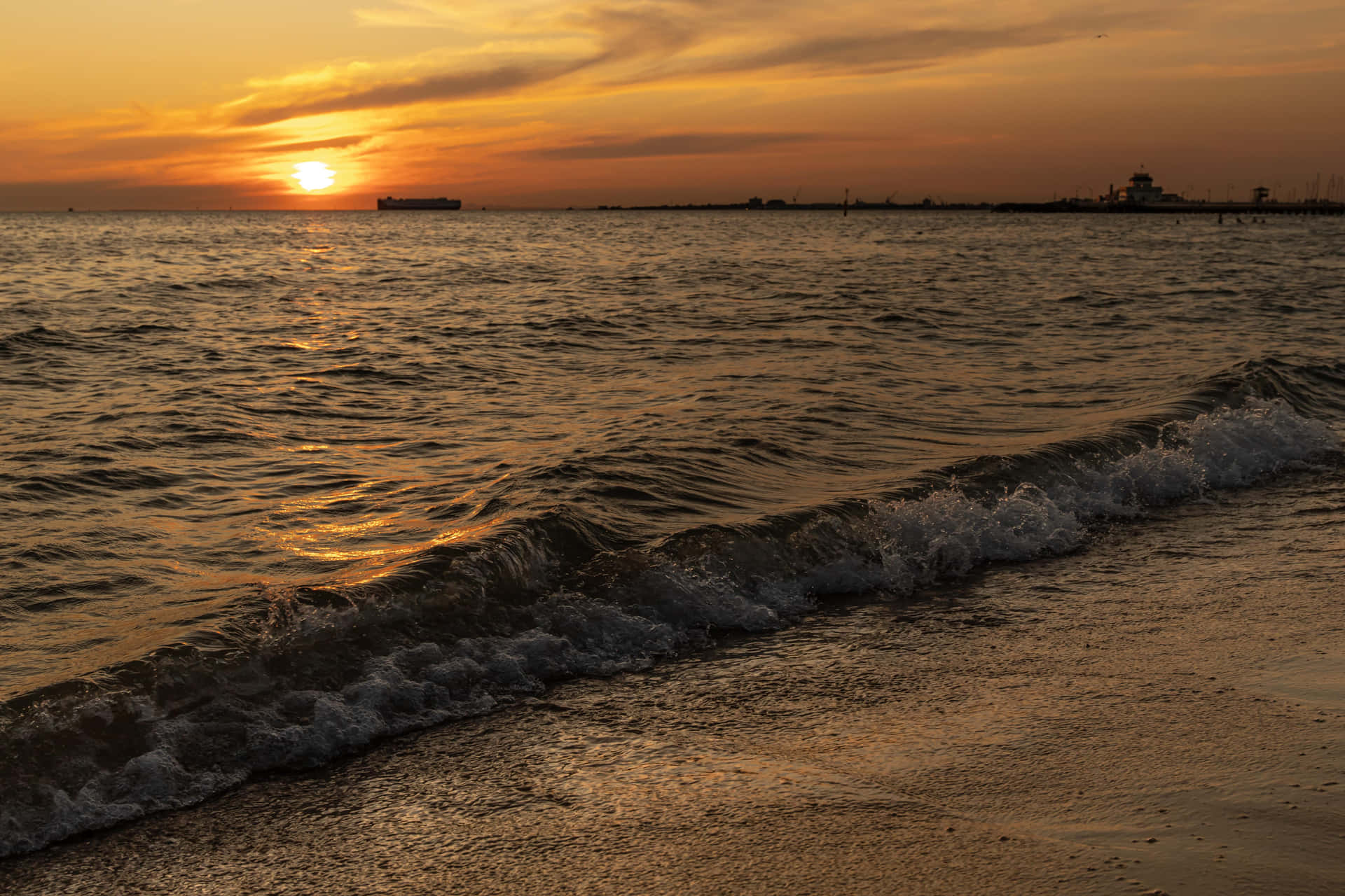 海洋上的夕陽，波浪沖刷海岸，遠處有船和燈塔。