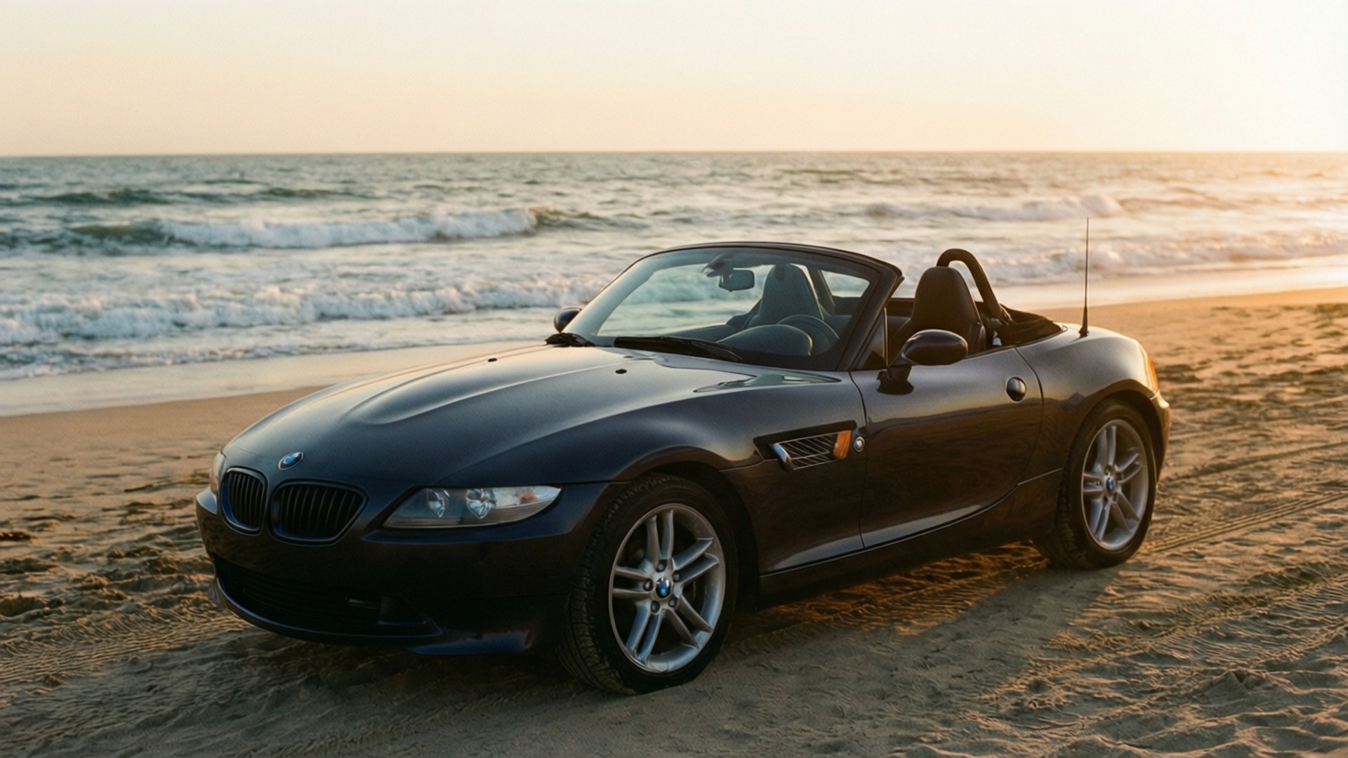 黑色BMW Z4開篷車停泊在沙灘上，夕陽下伴隨海浪。