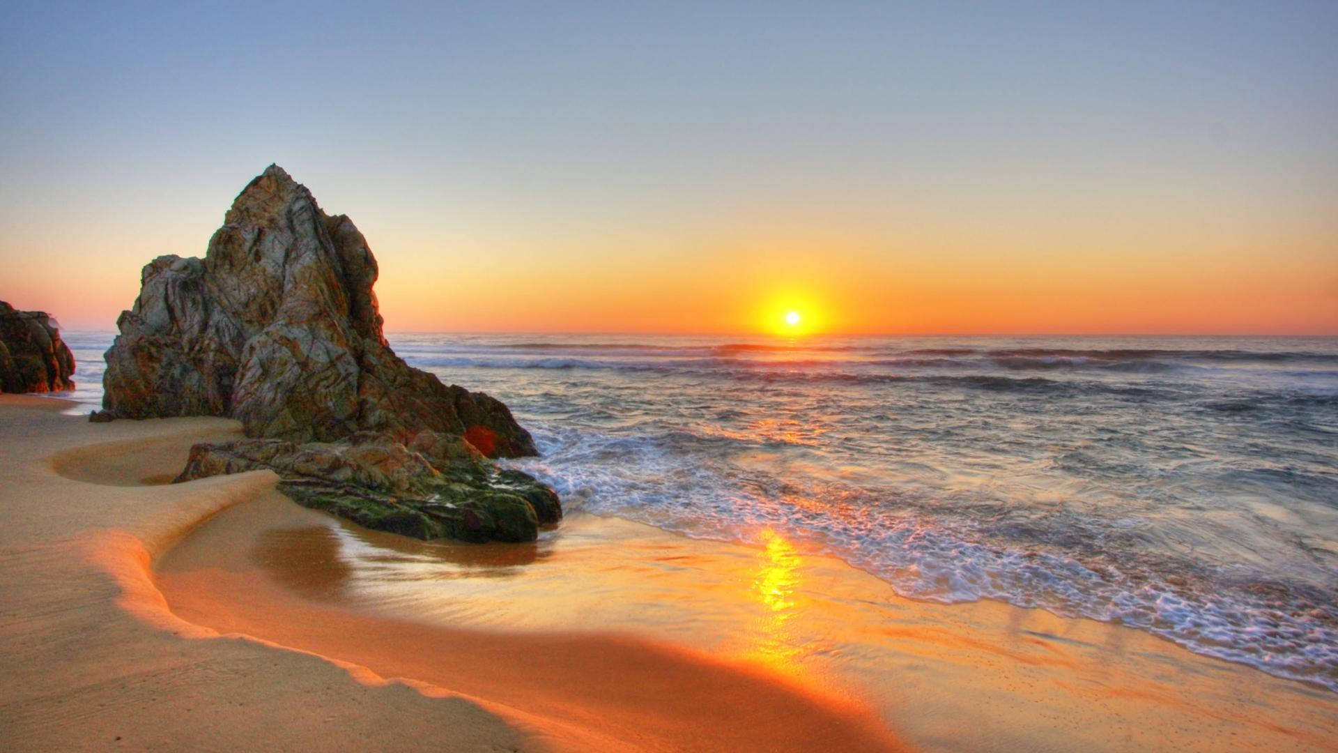 海灘日落時的巨大岩層 壁紙 — 夕陽將海洋和沙灘染成溫暖的橙色，海浪拍打著寧靜海灘上的一塊大岩石。