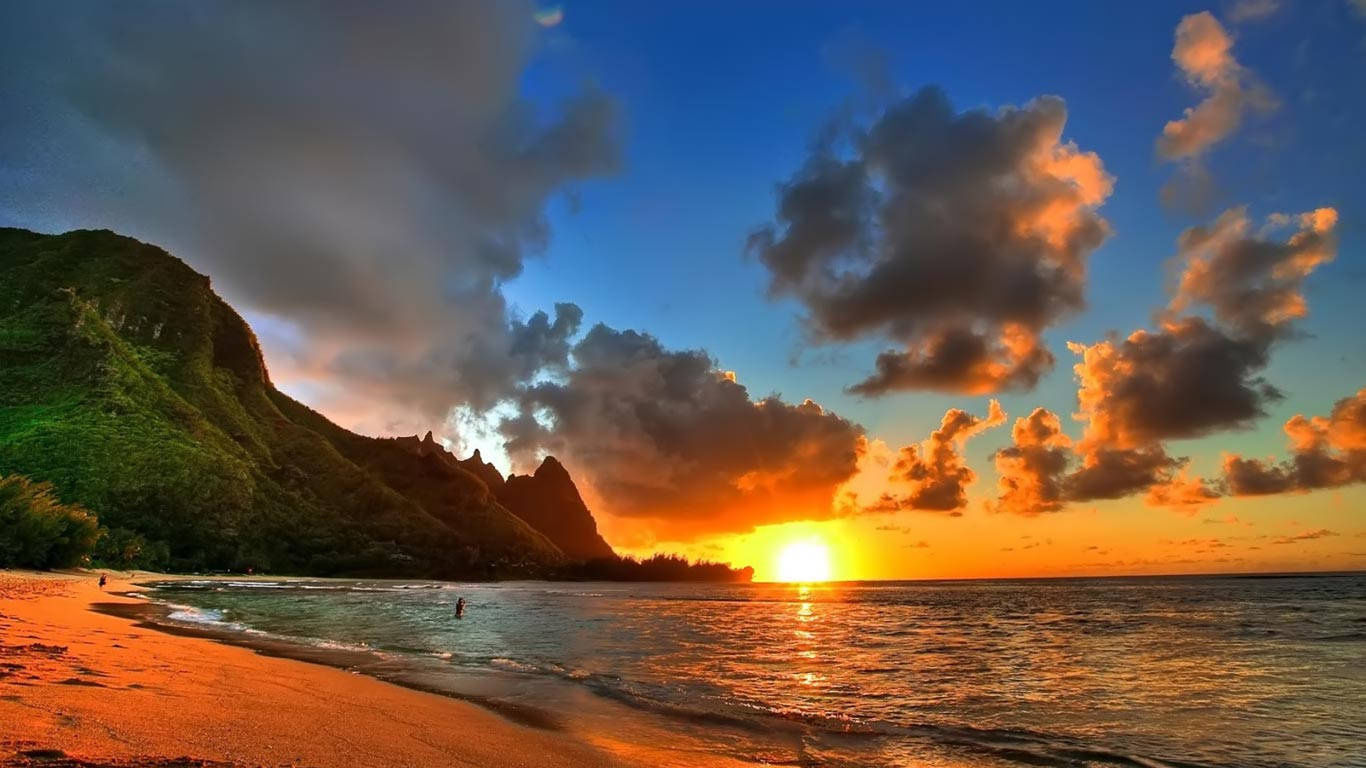海灘日落筆電桌面 壁紙 — 夕陽將天空染成橙色和藍色，海浪拍打沙灘，旁邊是鬱鬱蔥蔥的綠色懸崖。