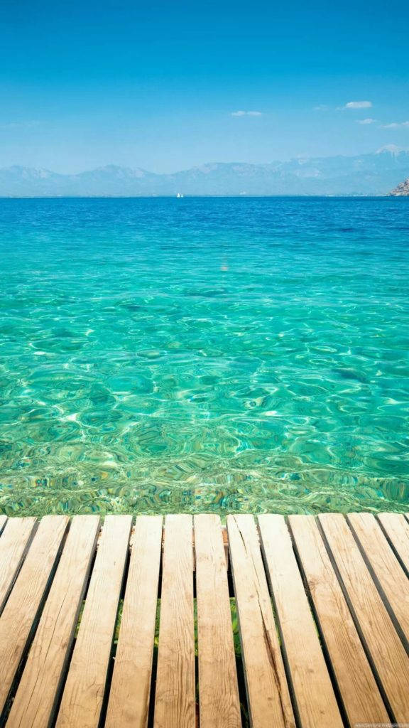 海灘木棧道 iPhone 壁紙 — 一個木製碼頭延伸到清澈的碧綠海水中，上方是明亮的藍天，遠處有山巒。