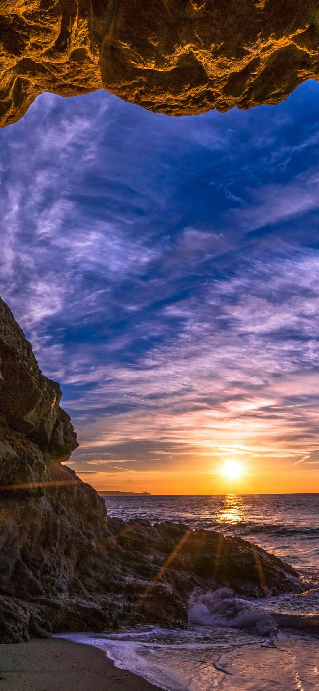 海灘洞穴與日落 Android 馬利布背景 壁紙 — 金色夕陽穿過岩石洞穴開口，照耀在波濤洶湧的海洋上，上方有紫色和藍色的雲彩。