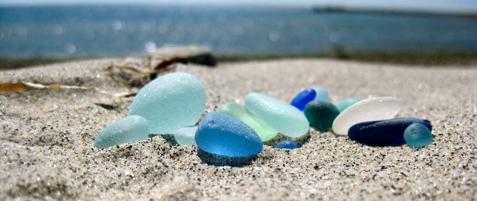 海灘海玻璃圖像 壁紙 — 光滑的海玻璃碎片，呈現藍色、綠色和白色色調，躺在陽光明媚的沙灘上，背景是模糊的海洋。