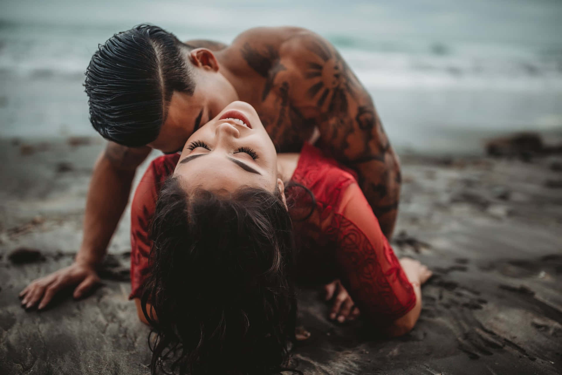 一個有紋身的男人在濕漉漉的海灘上親吻一個穿著紅色連衣裙微笑的女人。