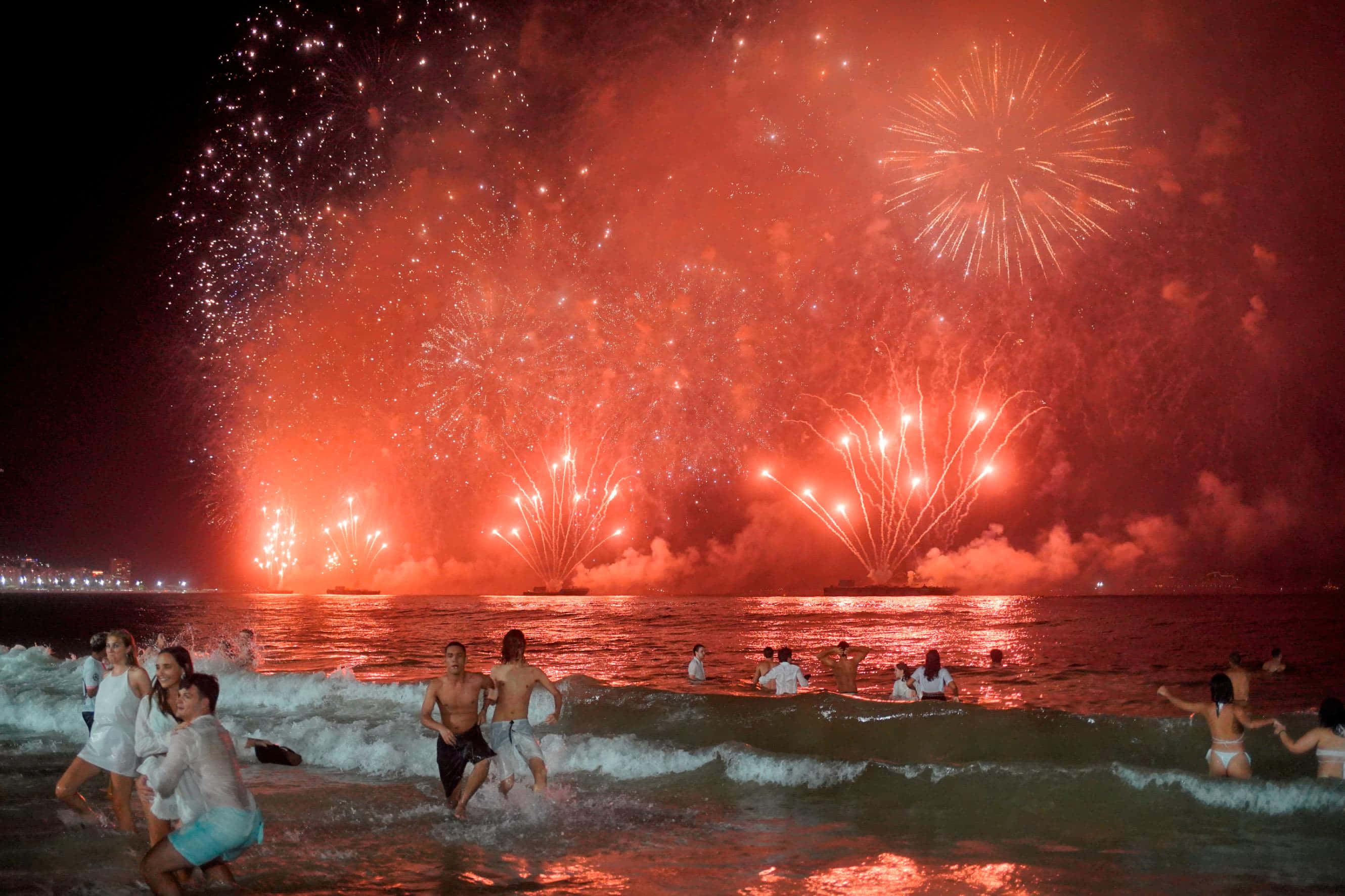 海灘煙火新年圖 壁紙 — 人們在海洋中慶祝，巨大的紅色煙火在夜間在水面上爆炸。