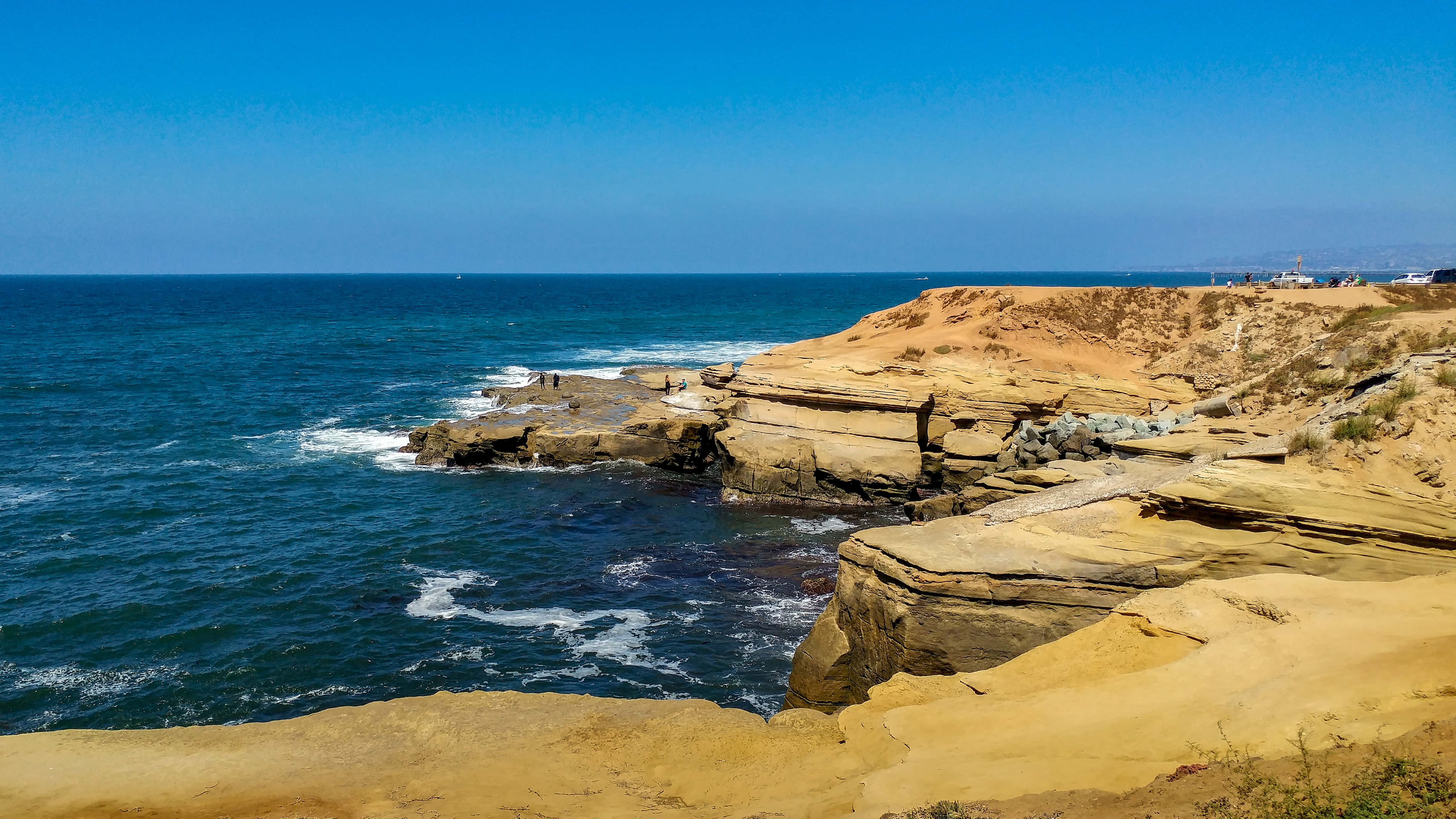 海灘的絕美風景 壁紙 — 陽光照射的金色懸崖與深藍色海浪在晴朗天空下撞擊層狀岩石結構相遇。