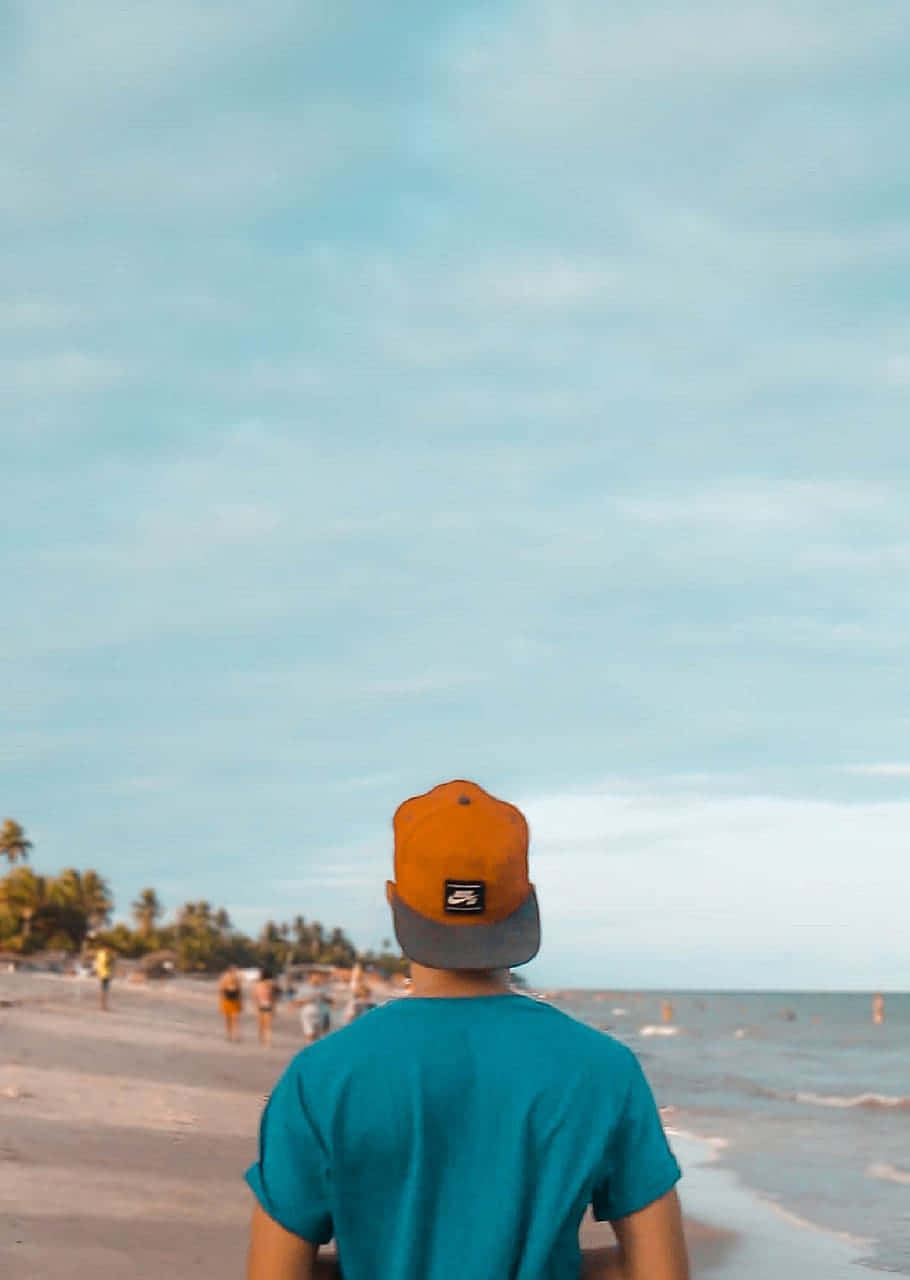 海灘美學旅行者圖片 壁紙 — 一個人穿著青綠色襯衫和橙色帽子，在柔和的藍天下沿著沙灘漫步，伴隨著溫柔的海浪。
