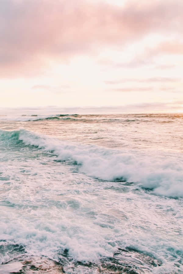 海灘美學海浪圖片 壁紙 — 粉紅和蜜桃色的雲彩在翻騰的海洋上發光，白色的泡沫波浪向岸邊滾動。