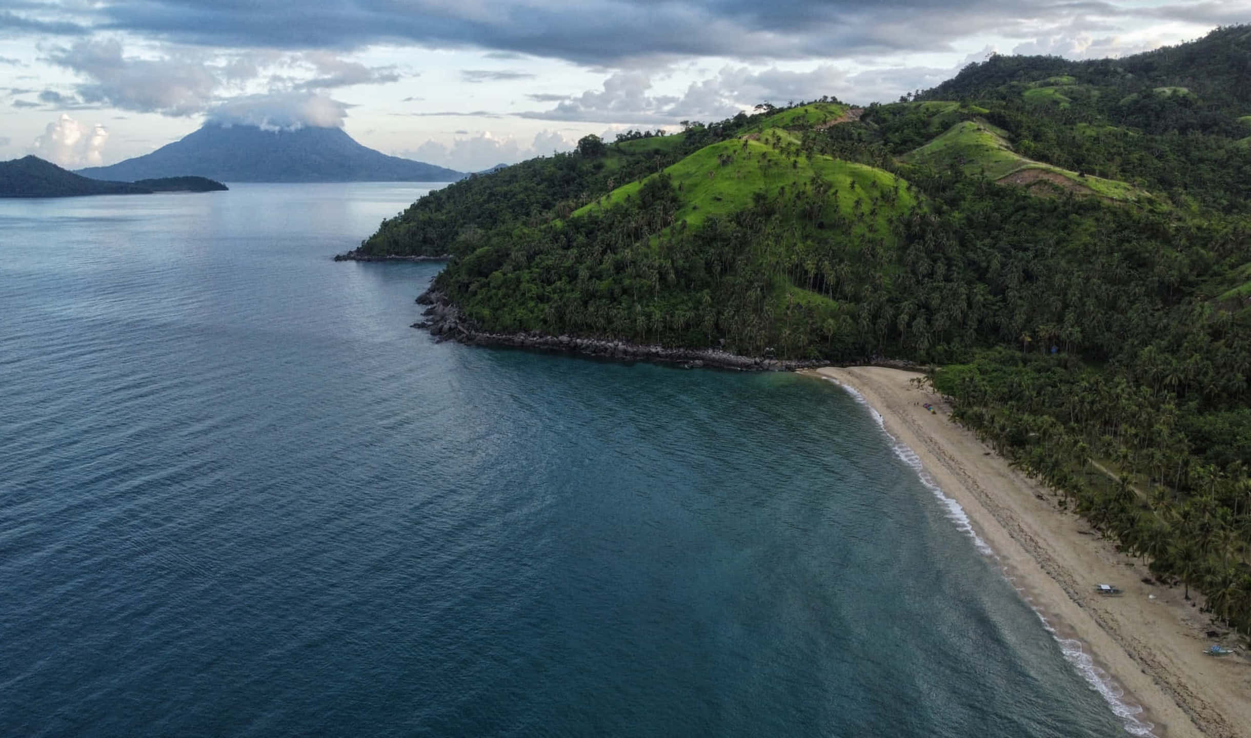 海灘與背景山脈 壁紙 — 鳥瞰圖：一個隱蔽的熱帶海灘沿著翠綠山丘蜿蜒，遠處有一座火山，天空多雲。