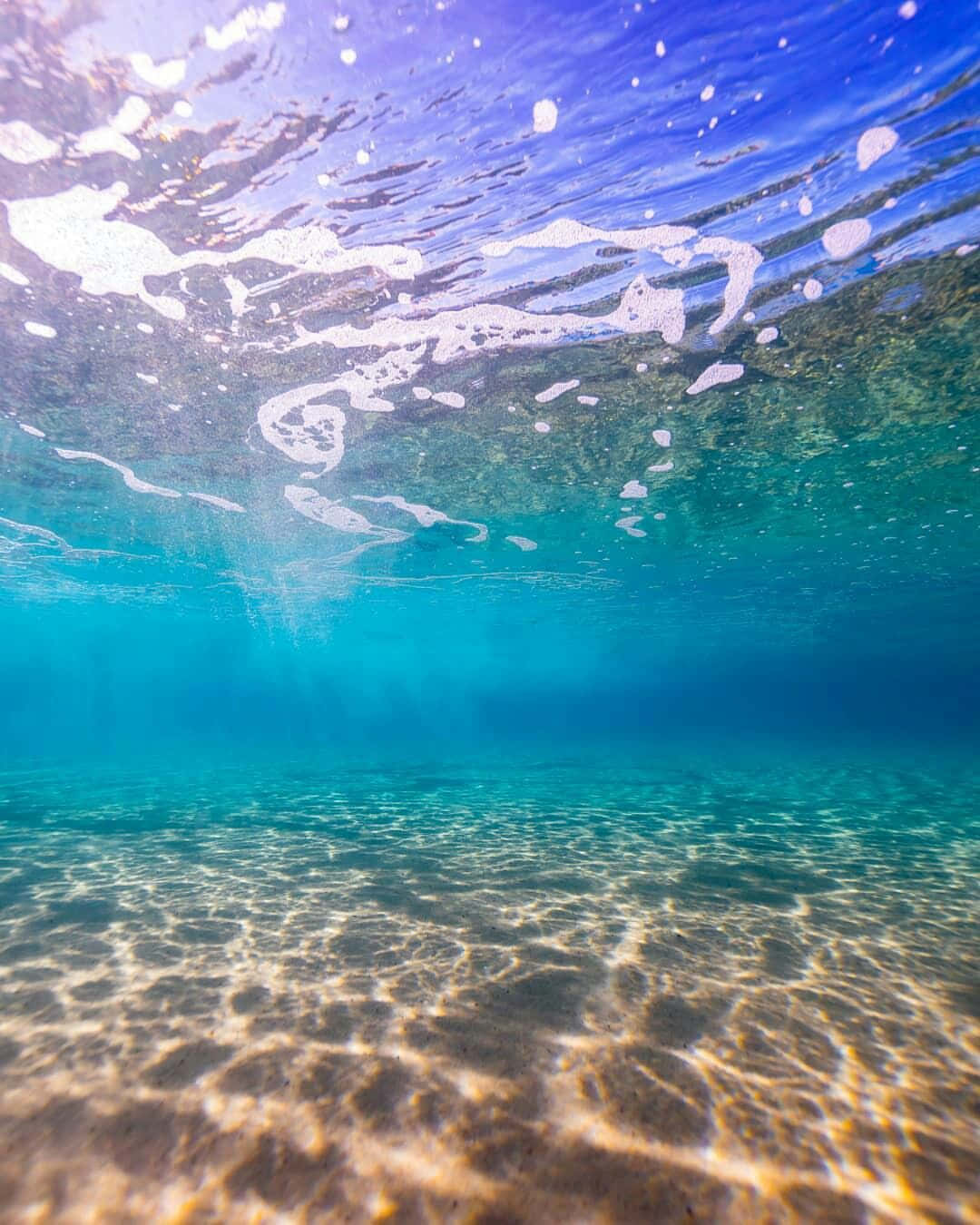 海灘陽光灑落海底圖 壁紙 — 陽光普照的碧綠海洋水下景觀，漣漪般的光影在沙質海底和上方泡沫表面舞動。
