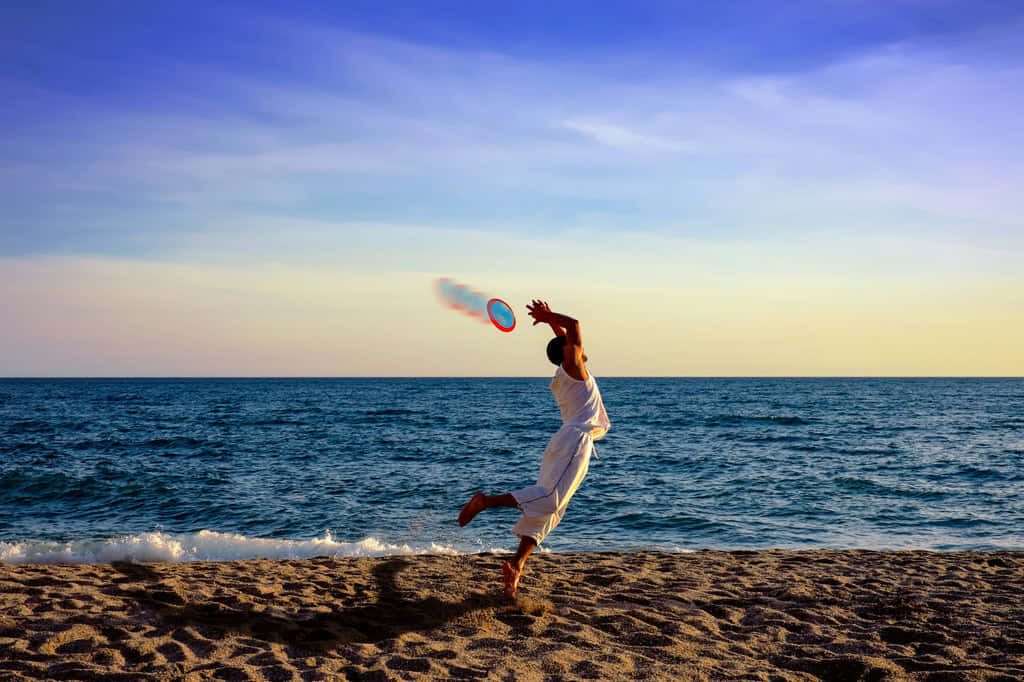 海灘風 Ultimate Frisbee 背景 壁紙 — 一個人在日落時分的沙灘上快樂地跳躍，在充滿活力的藍色和橙色天空下接住飛盤。