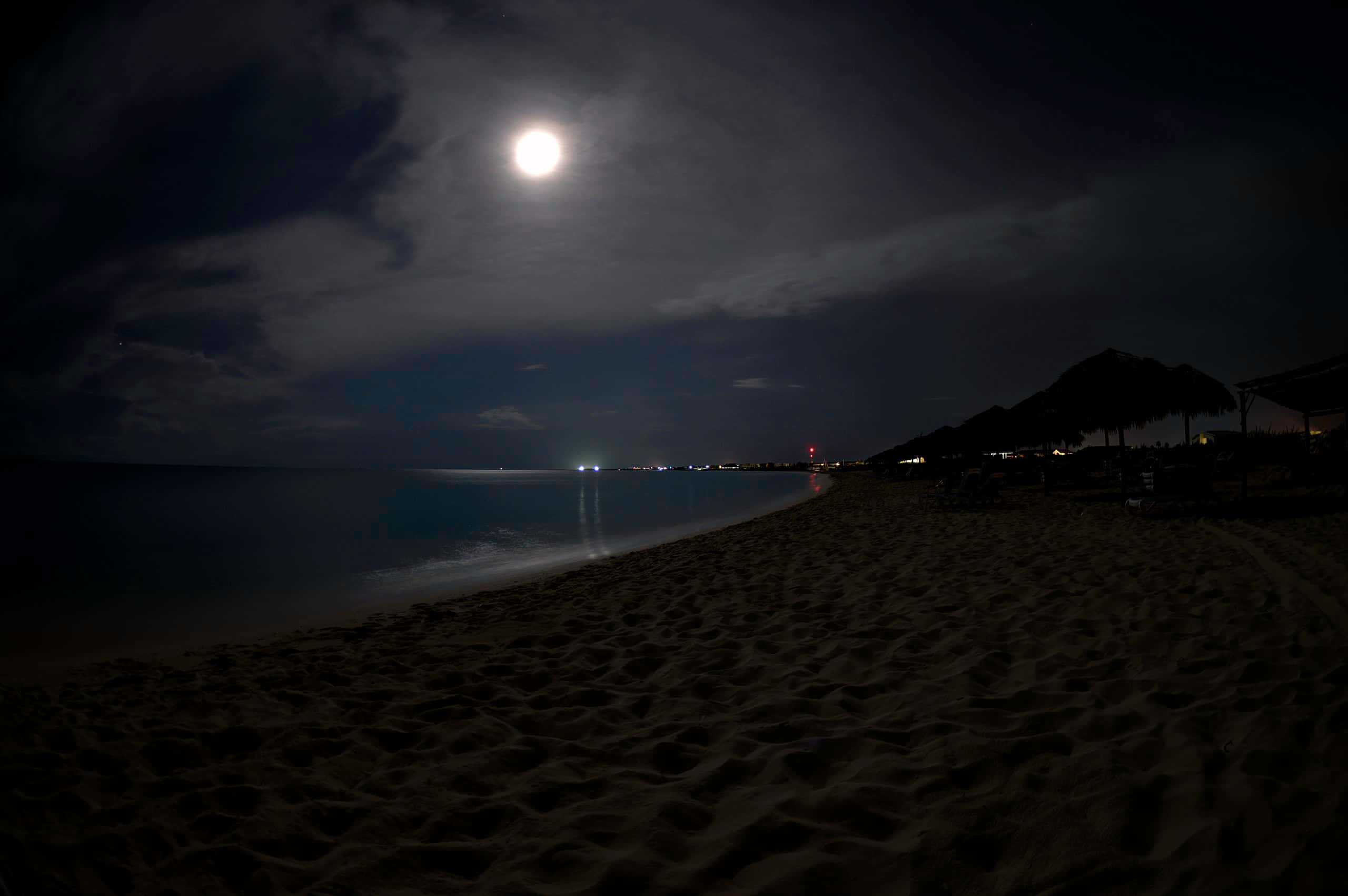 海灘魔幻夜景 壁紙 — 明亮的滿月在夜晚將銀色光芒灑在寧靜的海灘上，沙灘呈深色，地平線上有遠處的燈光。