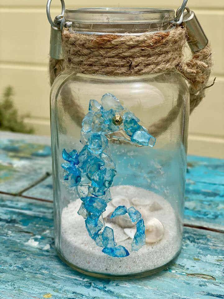 海玻璃海馬藝術項目圖 壁紙 — 一個帶麻繩蓋的玻璃罐，裡面裝著藍色水晶海馬和貝殼在沙中，放在風化的藍色木桌上。