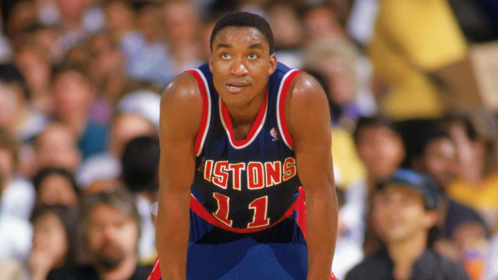 海耶斯·托馬斯1987年NBA賽季比賽 壁紙 — 一位身穿底特律活塞隊球衣的專注籃球運動員在比賽中向前傾，身後是模糊的人群。