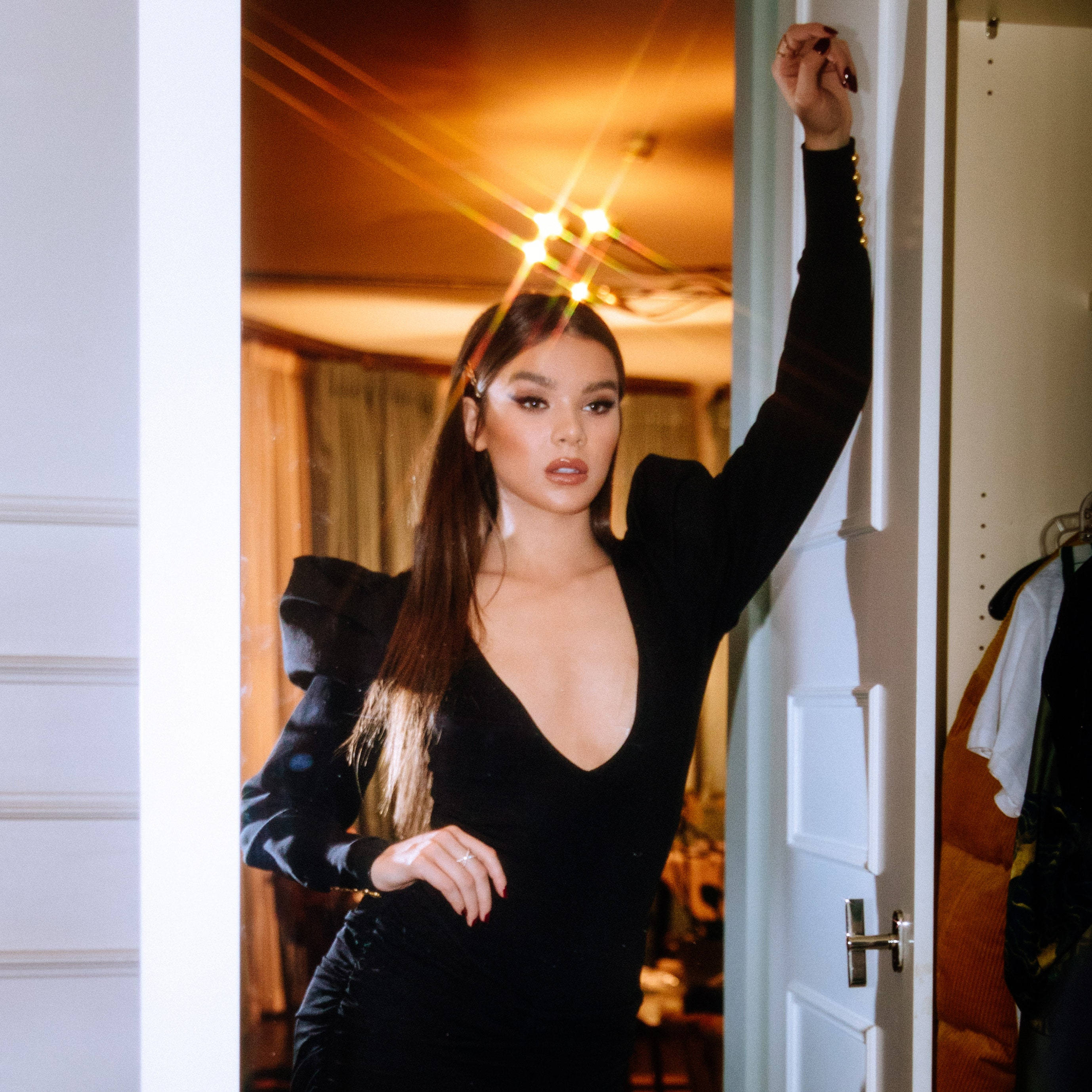 海莉·斯坦菲爾德《Vogue》 壁紙 — 一位身穿黑色天鵝絨連衣裙的女性自信地在門口擺姿勢，身後有溫暖的金色光芒閃耀。