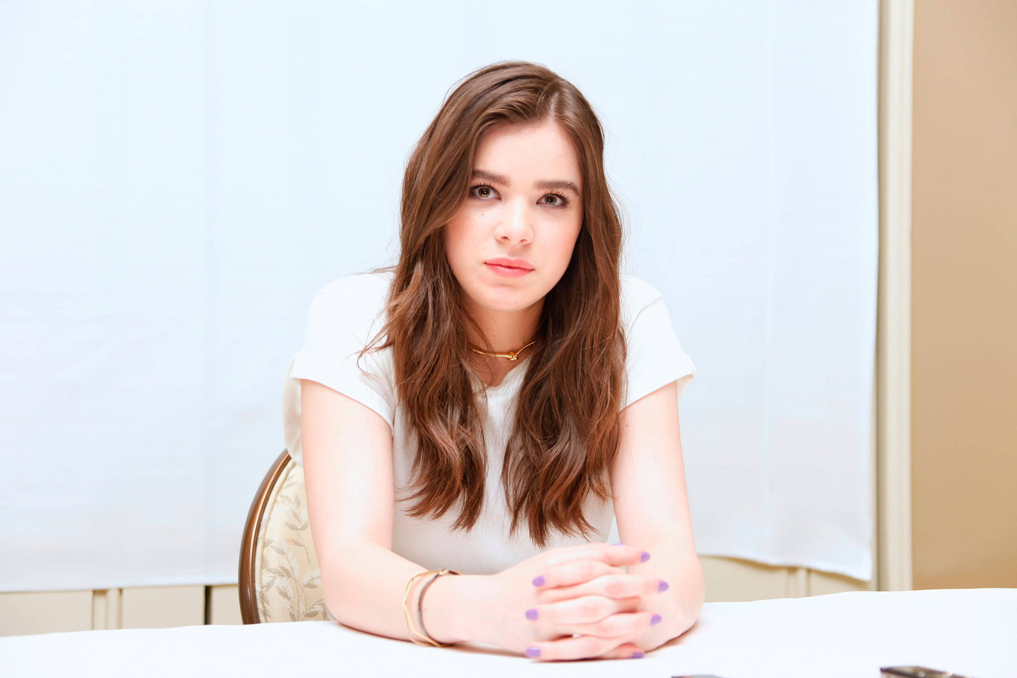 海莉·斯坦菲爾德出席《Pitch Perfect 2》記者會 壁紙 — 一位長棕色頭髮的年輕女性坐在白色桌子旁，穿著白色襯衫和紫色指甲油，直視鏡頭。