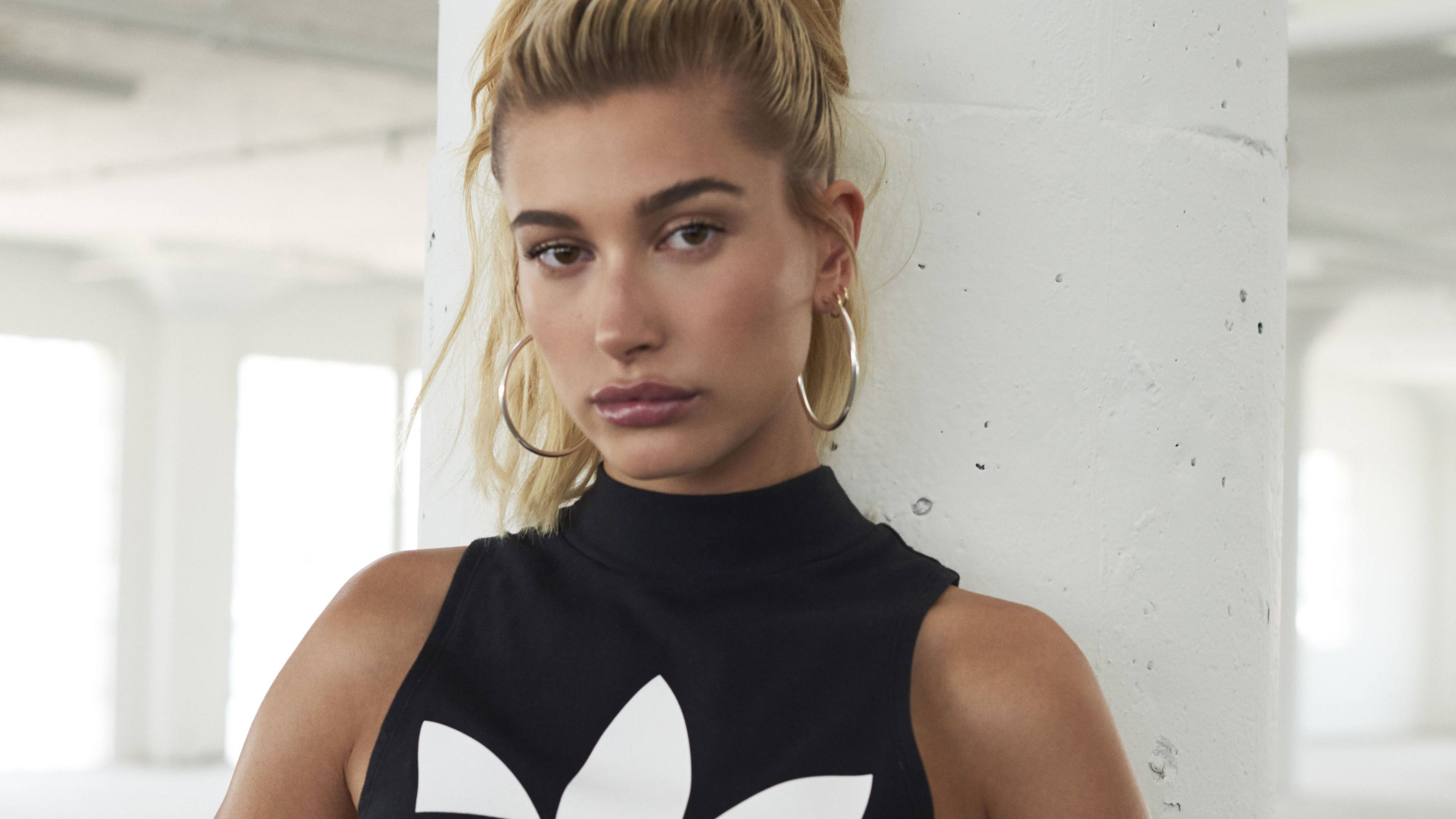 海莉·比伯為Adidas Originals拍攝 壁紙 — 一位金髮女子身穿帶有白色標誌的黑色無袖上衣，靠在明亮工業空間中的白色混凝土柱上。
