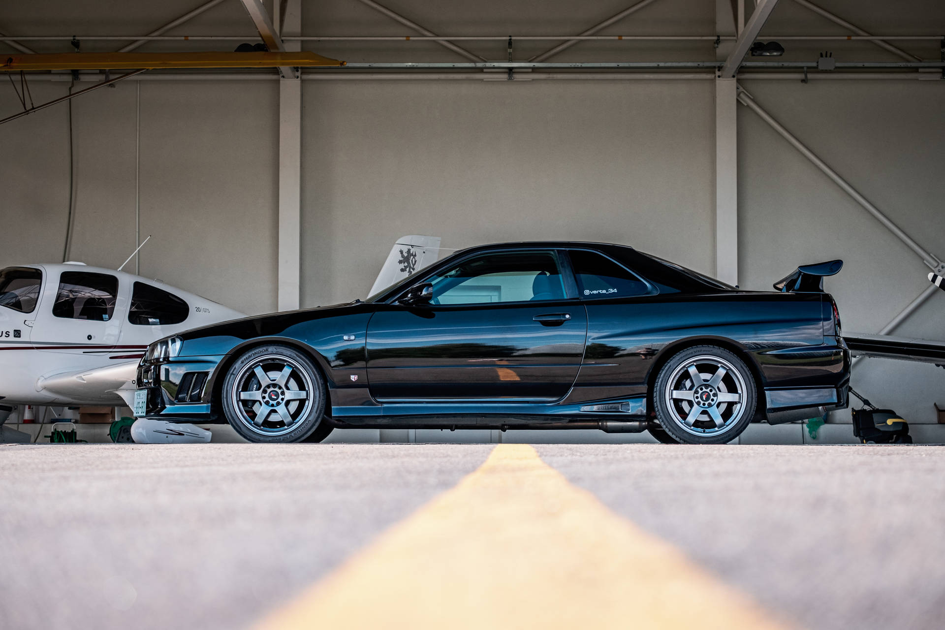 海藍色 Nissan Skyline GTR R34 壁紙 — 一架型格黑色日產Skyline GT-R停泊在機庫內，旁邊有一架小型白色飛機，並有一條黃色跑道線引向它。