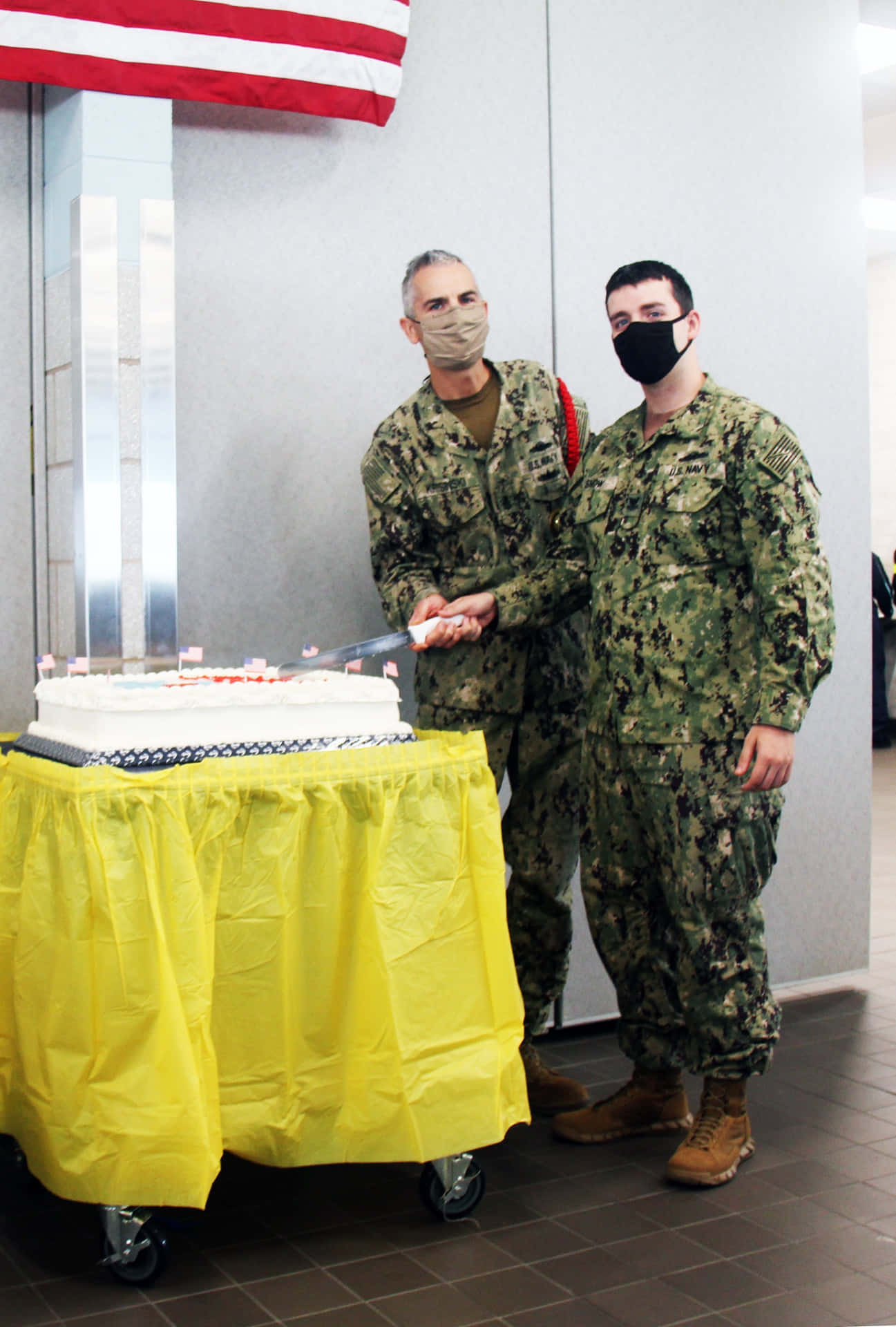海軍人員慶祝切蛋糕 壁紙 — 兩名戴著口罩、身穿迷彩軍服的軍人，在美國國旗下的一張黃色桌布上共同切一個大白色蛋糕。