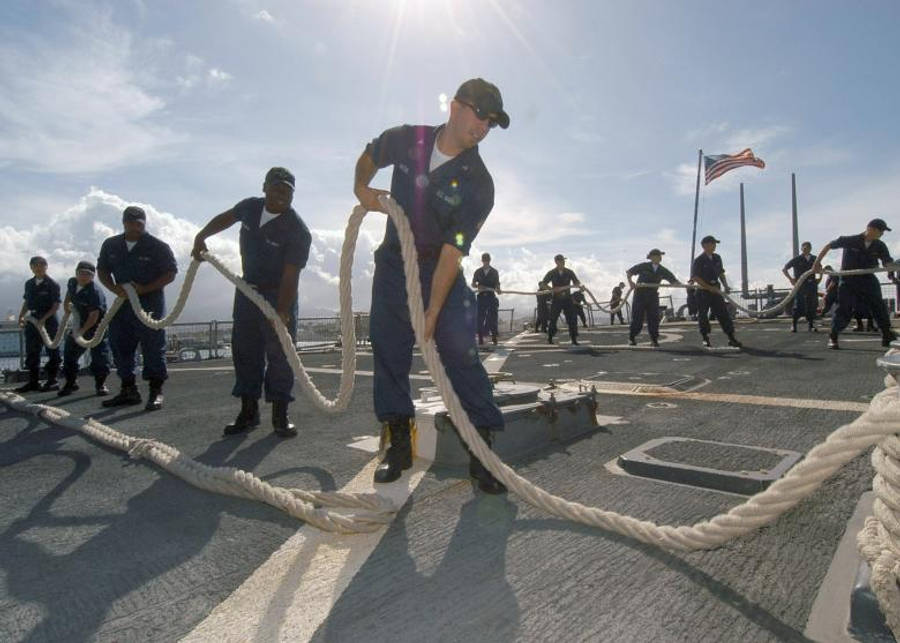 海軍士官齊心拉繩展現團隊合作 壁紙 — 身穿制服的海軍士兵在陽光明媚的甲板上用力拉著粗繩，美國國旗隨風飄揚。