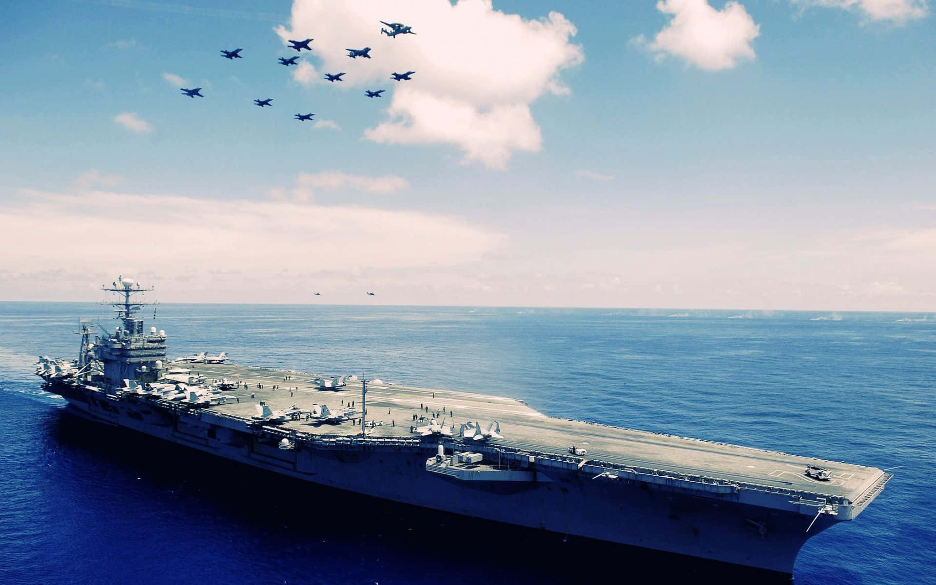 海軍官兵齊心為共同熱情而凝聚 壁紙 — 一艘巨大的航空母艦在深藍色的海洋中航行，天空明亮，戰鬥機在上方編隊飛行。
