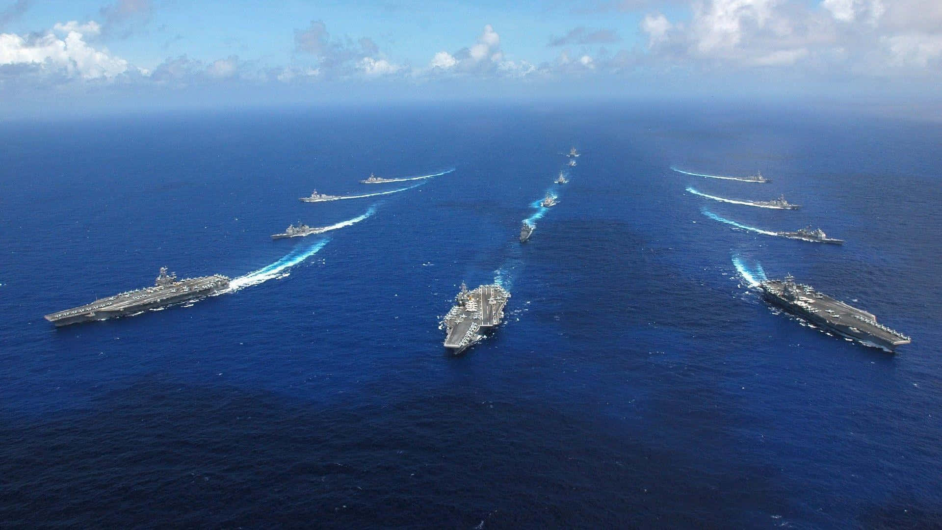 海軍戰艦準備起航 壁紙 — 多艘航空母艦和軍艦在部分多雲的天空下，於深藍色海洋上編隊巡航的鳥瞰圖。