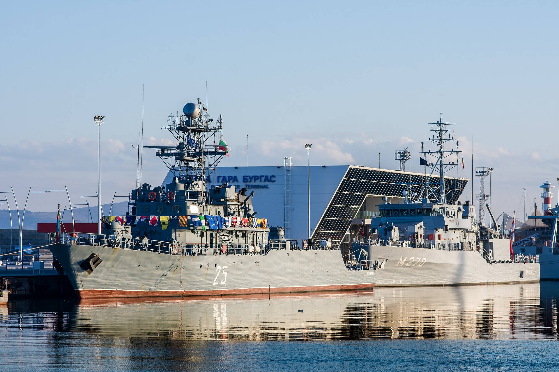 灰色海軍戰艦停泊在港口，後面有現代化的客運大樓。